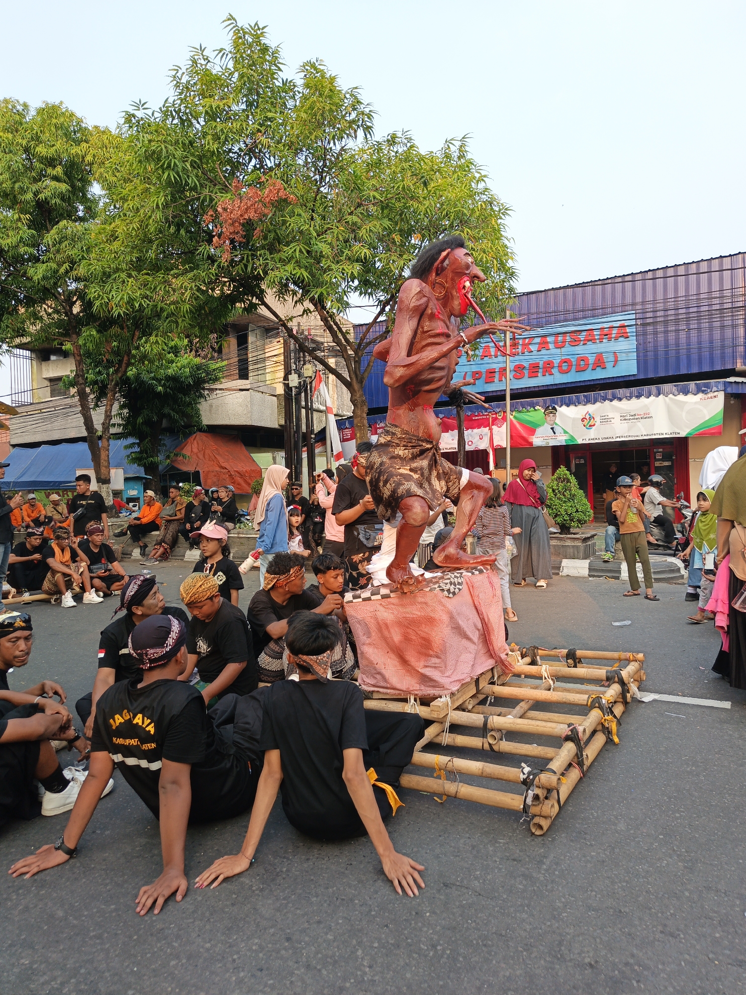 In Klaten, Indonesia, a large, brown, effigy of a grotesque figure with red accents stands on a bamboo platform in the middle of a street. The effigy is dressed in a patterned loincloth and appears to be part of a parade or festival. A crowd of people, many dressed in black shirts and some wearing traditional head coverings, are gathered around the platform. Some are seated on the ground, while others stand and observe. In the background, a building with a blue sign reading "ANEKA USAHA (PERSERODA)" is visible, along with trees and a clear sky. The overall scene suggests a community event or cultural celebration taking place outdoors in a public space.