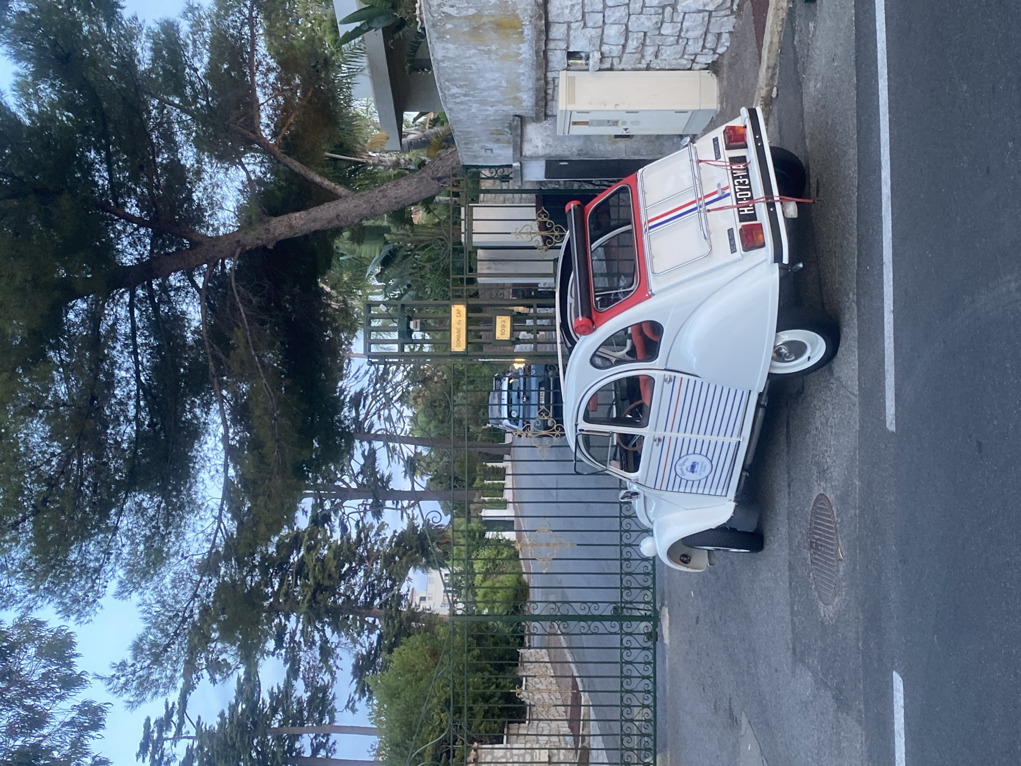 The image shows a white vintage Citroën 2CV parked on a street in Antibes, France. The car is decorated with blue and white stripes on its side and red, white, and blue stripes on the rear. It has a license plate that reads "H.J. 073-WA". Behind the car is a green metal gate leading to a property. Two signs are attached to the gate: one yellow sign reads "DOMAINE du CAP" and another yellow sign below it reads "1093". A dark blue SUV is visible through the gate. The street is paved and has white lane markings. In the background, there are trees and a building. The overall scene appears to be an exterior shot taken during the day, possibly in the late afternoon or early evening given the lighting.