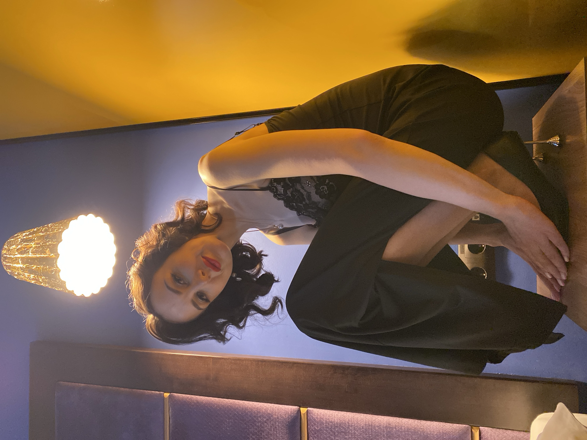 A woman with dark, curly hair and red lipstick is perched on the edge of a bed, facing the camera. She is wearing a black, lacy top and black pants. Her arms are wrapped around her legs, which are pulled up to her chest. The background consists of a purple headboard, a blue wall, and a yellow ceiling. A lamp is visible to the left of the woman. The location is Yekaterinburg, Russia.