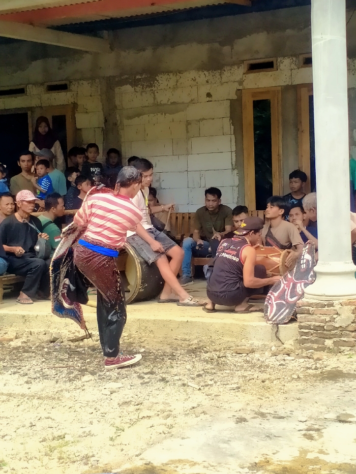 The image depicts an outdoor cultural performance in Ngargoyoso, Indonesia. In the foreground, a male performer wearing a red and white striped shirt, dark pants, and red sneakers stands on unpaved dirt ground. His pants appear wet, and he holds a large, dark, ornate prop with patterned and possibly feathered elements. To his right, on an elevated concrete platform, a male musician in a dark sleeveless shirt and a beret plays a light brown traditional drum (kendang) while in a squatting position. A decorative, wing-shaped object with red and white patterns rests on the platform beside him. Another large, dark drum is visible behind the musician. Numerous spectators, including men, women, and children, are seated or standing on the platform and within the doorways of an unfinished concrete block building, observing the event. The building features exposed light-colored concrete blocks and wooden doors, topped with a reddish-brown corrugated metal roof.