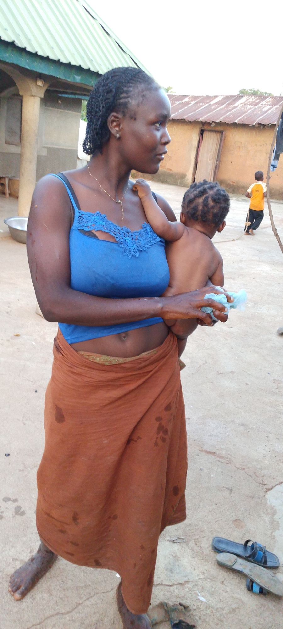 In a courtyard, a woman of color is holding a naked baby. The woman wears a blue tank top and an orange skirt. The baby is facing away from the camera, being held on the woman's left side. The woman holds a blue sponge or cloth in her right hand. In the background, a young child in a yellow shirt and dark pants is walking away from the camera. The environment appears to be rural, with simple buildings and an unpaved ground surface. The location is Kamantan, Nigeria.
