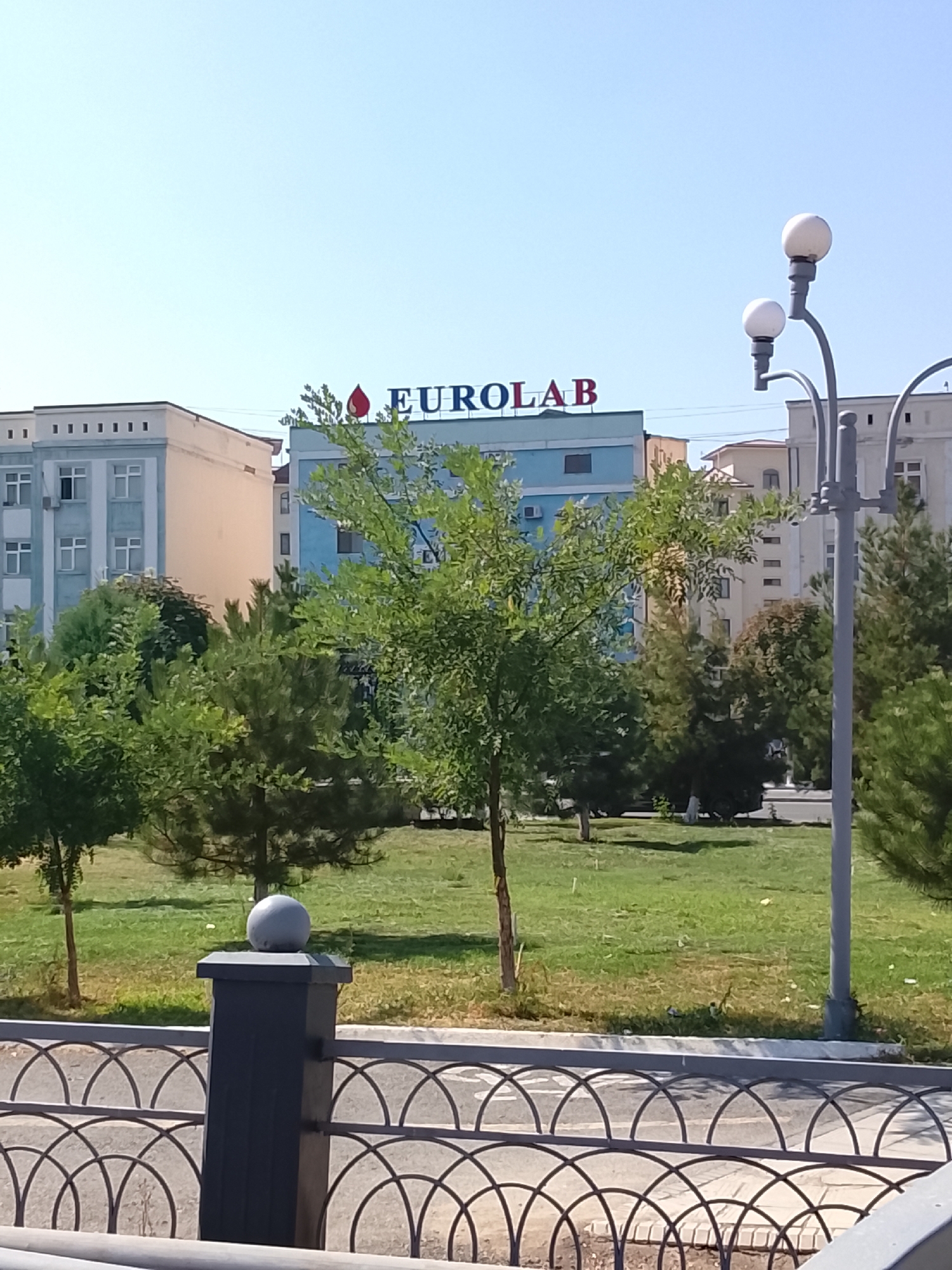 The image depicts a bright, sunny day in Fergana City, Uzbekistan, showcasing a multi-story building with a prominent "EUOLAB" sign on its facade. The building appears to be a medical facility or laboratory, suggested by the sign and the presence of trees and a manicured lawn in front. A decorative lamppost with two globes stands to the right, and a metal fence with an ornate design runs along the bottom of the frame. Other buildings, some lighter in color and with numerous windows, are visible in the background, suggesting an urban or suburban setting. The sky is clear and blue, indicating good weather conditions. There are no people visible in the image.
