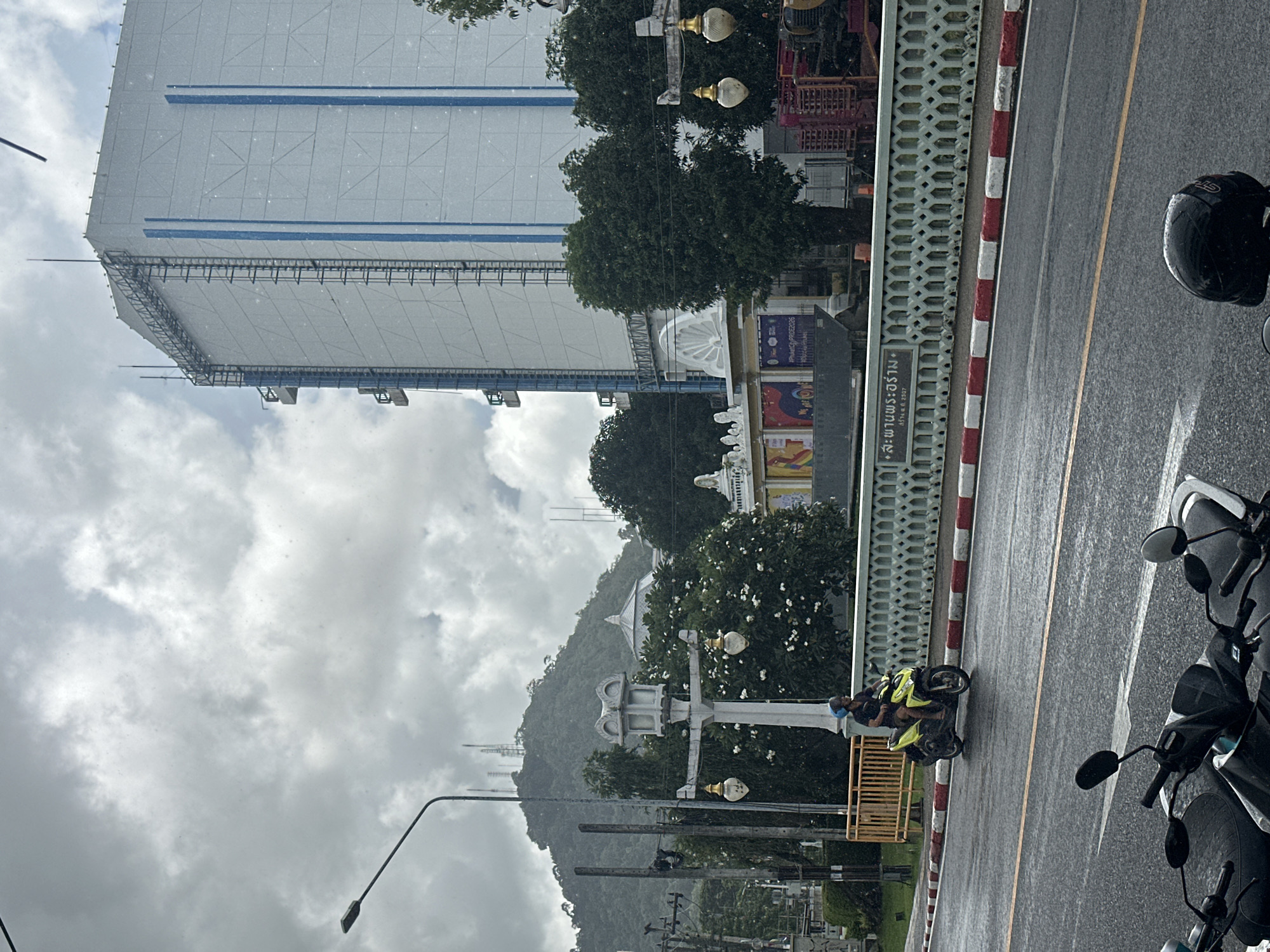 The image depicts a street scene in Phuket City Municipality, Thailand, under overcast and rainy conditions. The perspective is from a moving vehicle, likely a motorcycle, given the presence of other motorcycles in the foreground and a rider on a motorcycle in the mid-ground.
The scene features a large, modern building with a white facade and blue accents dominating the background, appearing to be under construction or renovation, covered in protective sheeting. To the right of this building, trees and a glimpse of a dome-like structure suggest a more established area, possibly a temple or public building. A mountainous landscape is visible further in the distance, shrouded in mist.
A distinct white and red striped barrier runs alongside the road, separating it from a sidewalk or pedestrian area. On the road itself, traffic markings are visible, indicating lanes for vehicles. The road surface is wet, consistent with the rainy weather.
A person on a motorcycle, wearing a yellow reflective vest and a blue helmet, is the most prominent human subject, traveling on the road. Other motorcycles are parked or moving in the foreground, suggesting a busy urban environment.
Visible text includes a sign on the barrier that reads "สะพานพระรัง" and below it "เริ่ม ต.ค. 2557" (meaning "Phra Rang Bridge" and "Started Oct. 2014"). Other signs with Thai script and some English text are partially visible on buildings in the mid-ground, but are too blurry to read clearly.
The overall mood is somber due to the cloudy and wet weather, but the scene conveys a sense of everyday urban activity in Thailand. The time of day appears to be daytime, but the overcast sky limits the direct sunlight.