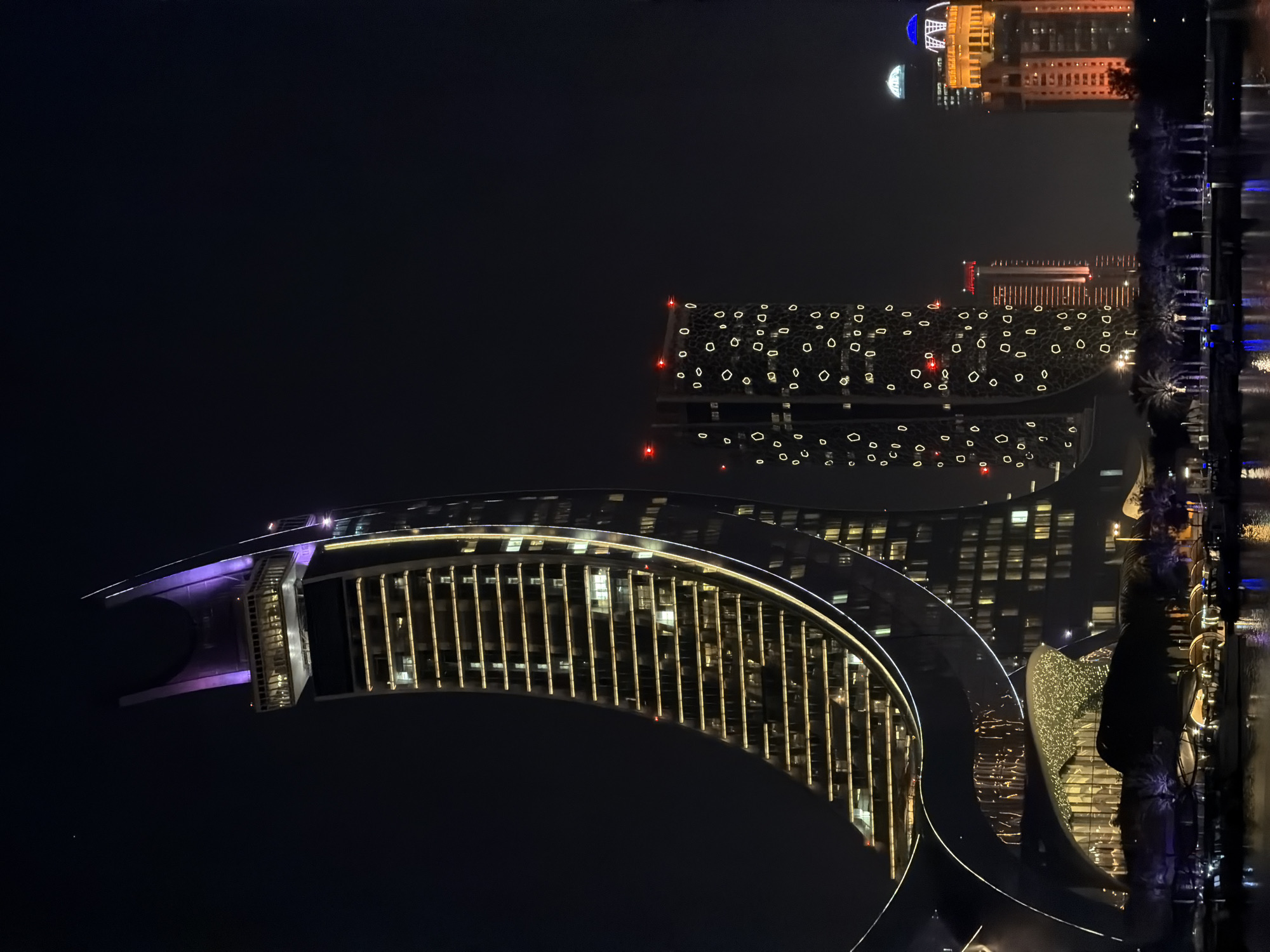 This media file is a nighttime photograph of a modern architectural scene. The primary subjects are several large, illuminated buildings reflecting in what appears to be a body of water. The building in the foreground is a large, curved structure with a significant portion covered in vertical, warm-toned lights, resembling a ribbed facade. A section of this building is lit with purple light. Behind it, another building has a darker facade punctuated by numerous circular and diamond-shaped openings, each illuminated with a soft yellow light. Further in the distance, taller buildings are visible, one with a distinctive golden-colored structure and a blue dome-like feature at its apex, and another with a grid of warm lights.
The scene is set at night, as indicated by the darkness of the sky and the artificial illumination of the buildings. There are reflections of the lights on the water's surface, suggesting a calm water body, possibly a harbor or a canal, adjacent to the buildings. Palm trees are visible along the waterfront, adding a tropical or sub-tropical cue to the location. The overall mood is one of urban sophistication and modern design. No people are discernible in the image, and no text is visible. The time of day is clearly night. The weather appears clear, allowing for the reflections on the water.