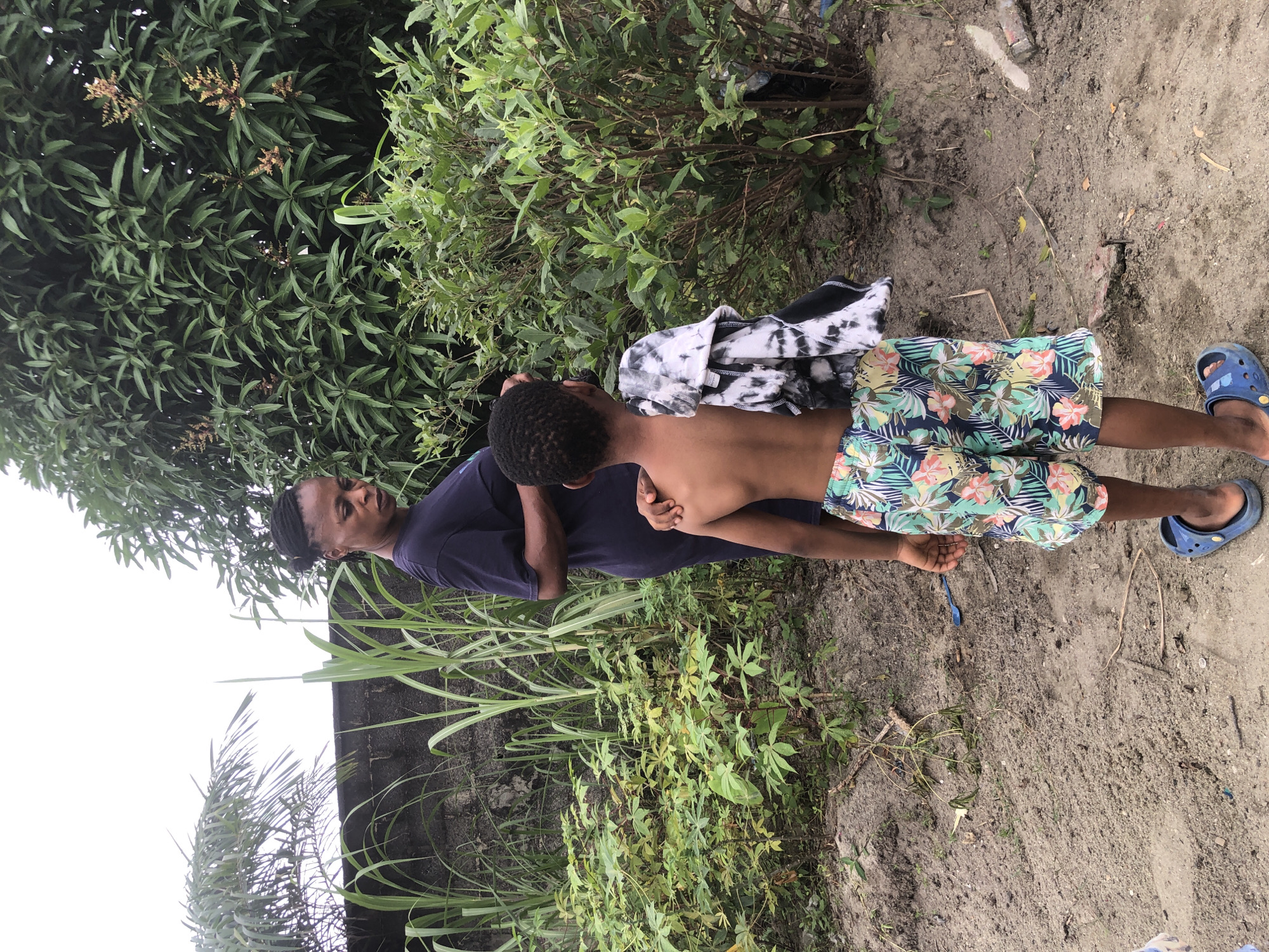 In Ado, Nigeria, a young Black boy, facing away from the camera, stands in the foreground. He is wearing patterned shorts and blue Crocs. A tie-dye t-shirt is draped over his left shoulder. He appears to be interacting with a Black woman, seen from the shoulders down, wearing a dark blue t-shirt and also facing away from the camera. She has her left hand on the boy's shoulder. They are standing in a dirt-covered area with sparse vegetation. Behind them, there is a dense green bush, a wall, and tall leafy plants.