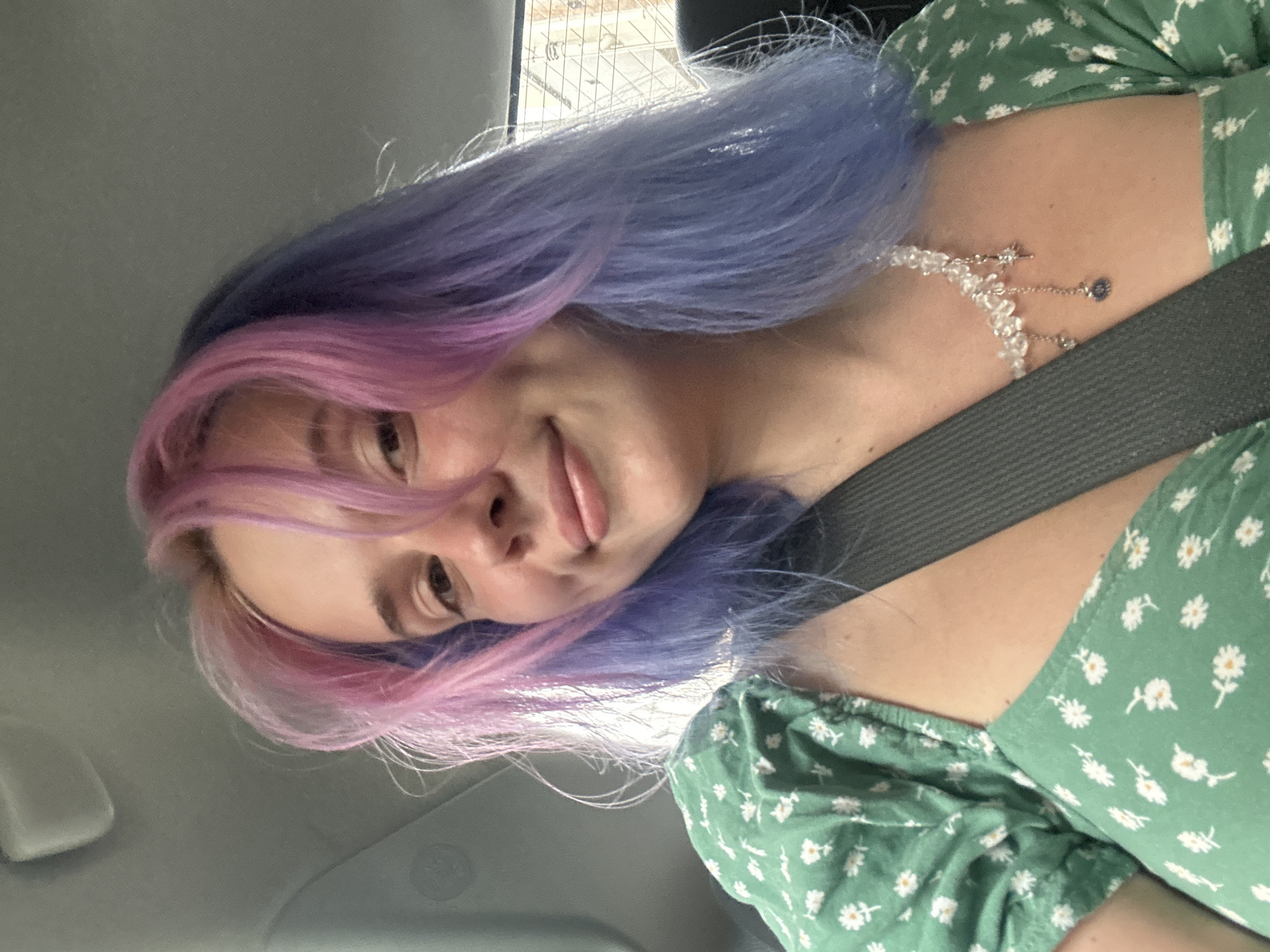 A selfie-style photograph shows a person with two-toned hair (pink and blue) wearing a green floral dress and a pearl-like necklace, positioned inside a vehicle with a grey seatbelt. The photo appears to be taken from a low angle, angled upwards towards the person's face. The surroundings are partially visible through the vehicle's window, suggesting an urban setting; however, without more specific visual indicators, determining whether this image was taken in Gijón/Xixón, Spain is impossible.