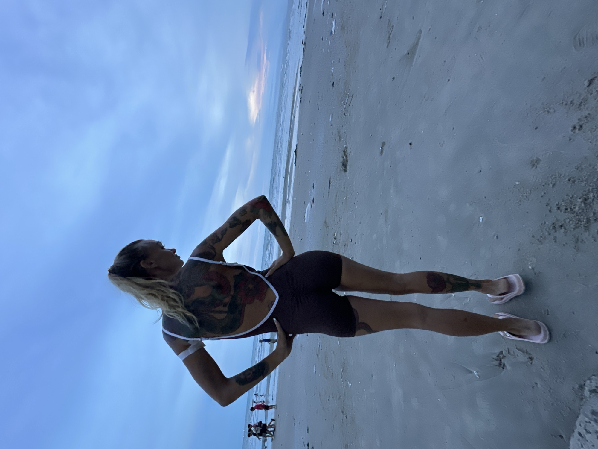 A woman with extensive tattoos on her back, arms, and legs stands on a sandy beach, facing away from the camera. She is wearing a dark brown, one-piece bathing suit with white trim and light pink flip-flops. Her hands are on her hips, and her body is angled slightly. The background shows a cloudy sky, the ocean, and a stretch of wet sand. In the distance, several other people are visible walking along the shoreline. The lighting suggests it is either early morning or late evening.