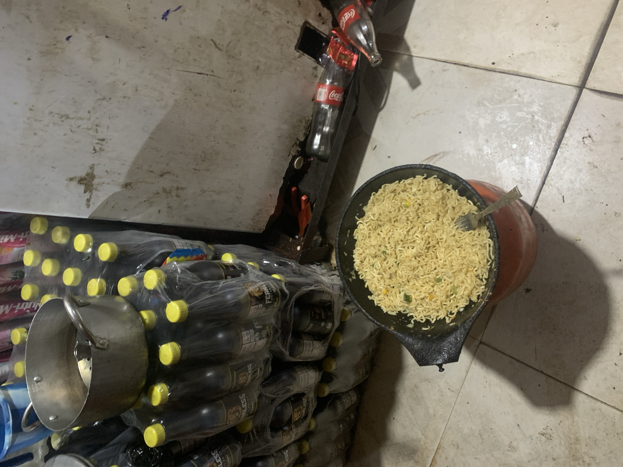 The image depicts an indoor scene, likely in a commercial or storage area, with stacks of bottled drinks and a pot of noodles. In the foreground, a black pot filled with cooked instant noodles sits on a red base on a tiled floor. A fork rests on the edge of the pot, partially submerged in the noodles. Behind the pot, several stacks of plastic-bottled drinks, predominantly dark-colored with yellow caps, are visible. These bottles appear to be branded with labels, some showing the word "FEARLESS." In the background, a silver pot with handles is placed on top of one of the stacks of bottles. To the right, a white, stained surface, possibly a wall or refrigerator, is visible, with some discarded Coca-Cola bottles and a black object resembling a smartphone lying on a shelf or ledge. The overall environment appears somewhat cluttered and possibly indicative of a storage or preparation area. The scene does not provide specific visual cues to pinpoint the exact location within the city of Jos, Nigeria, beyond it being an indoor setting.