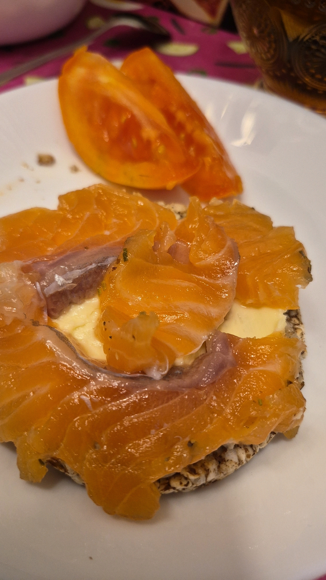 A close-up, overhead shot shows a plate of food. In the foreground, a rice cake is topped with a spread of yellow butter, slices of smoked salmon, and thin slivers of red onion. To the upper right of the rice cake, two wedges of bright orange tomato are visible. The food is presented on a white, circular plate. The background is blurred, showing hints of a pink and green patterned tablecloth and a dark glass. The scene is illuminated by a warm light source. No people or specific location in Krauja, Latvia are visible.