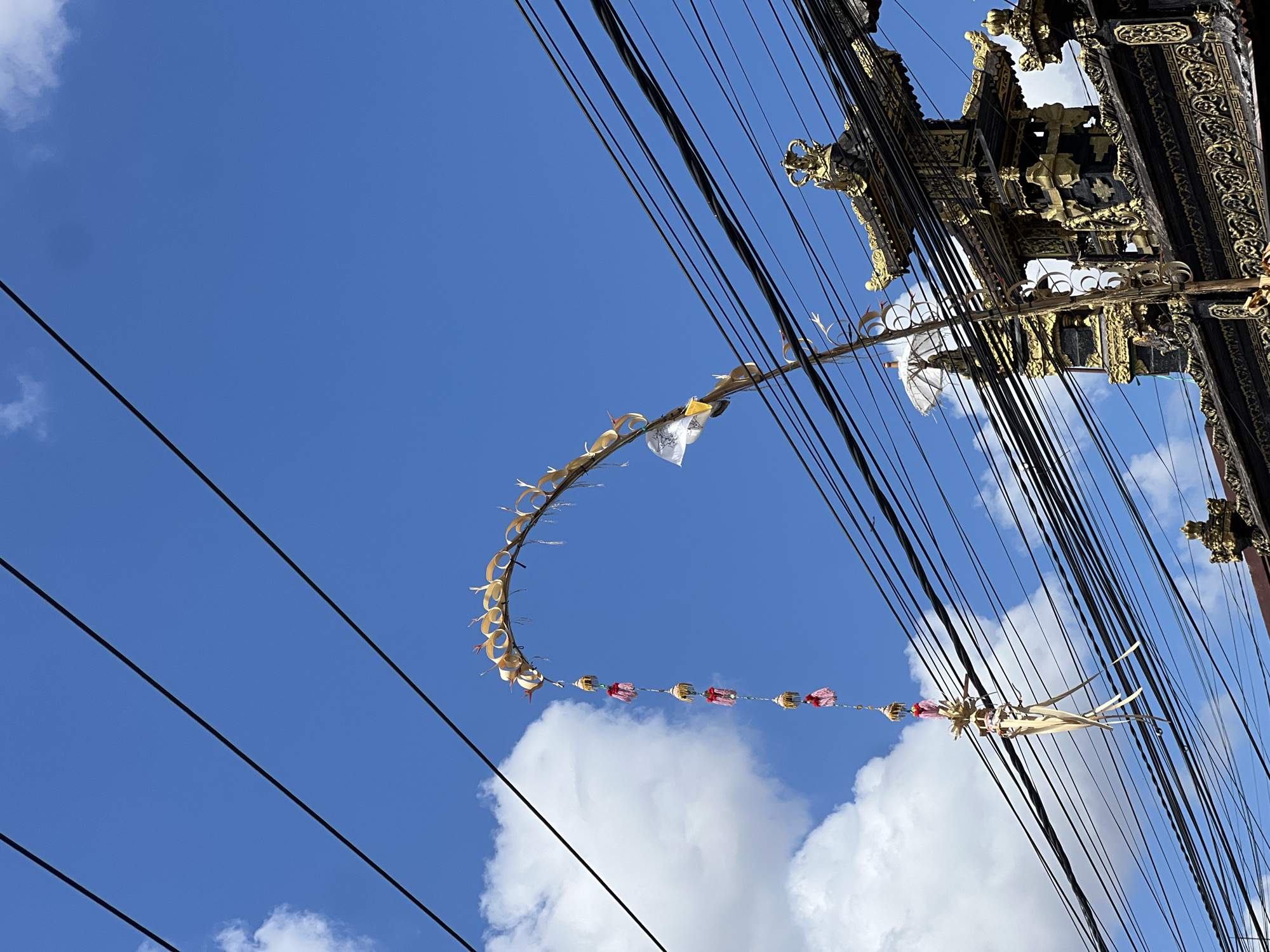 In Denpasar, Indonesia, an outdoor scene unfolds beneath a vibrant blue sky dotted with scattered white clouds. Overhead, a dense network of black electrical wires stretches across the frame. A decorative, arched banner, made of woven palm leaves and adorned with red and gold ornaments, hangs suspended from these wires. Below the banner, a tall, slender wooden pole extends downwards, also decorated with similar ornaments and palm leaf elements. In the background, partially obscured by the wires, is the ornate facade of a traditional Balinese building. This structure features intricate black and gold detailing, including tiered roofs and decorative carvings, suggesting it is a temple or a significant cultural site. The overall impression is a snapshot of urban Balinese life, where traditional decorations are integrated into the modern infrastructure of overhead wiring.