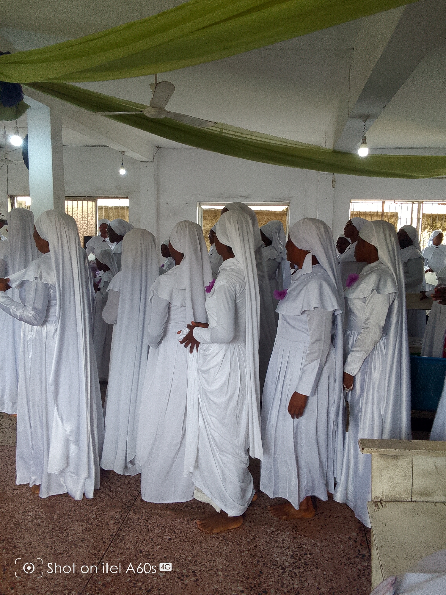 A group of people in white robes and head coverings stand indoors. The robes are long and flowing. Some of the individuals have bare feet. The interior is decorated with green fabric draped from the ceiling. There is a ceiling fan and light fixtures visible. The background is blurry.
