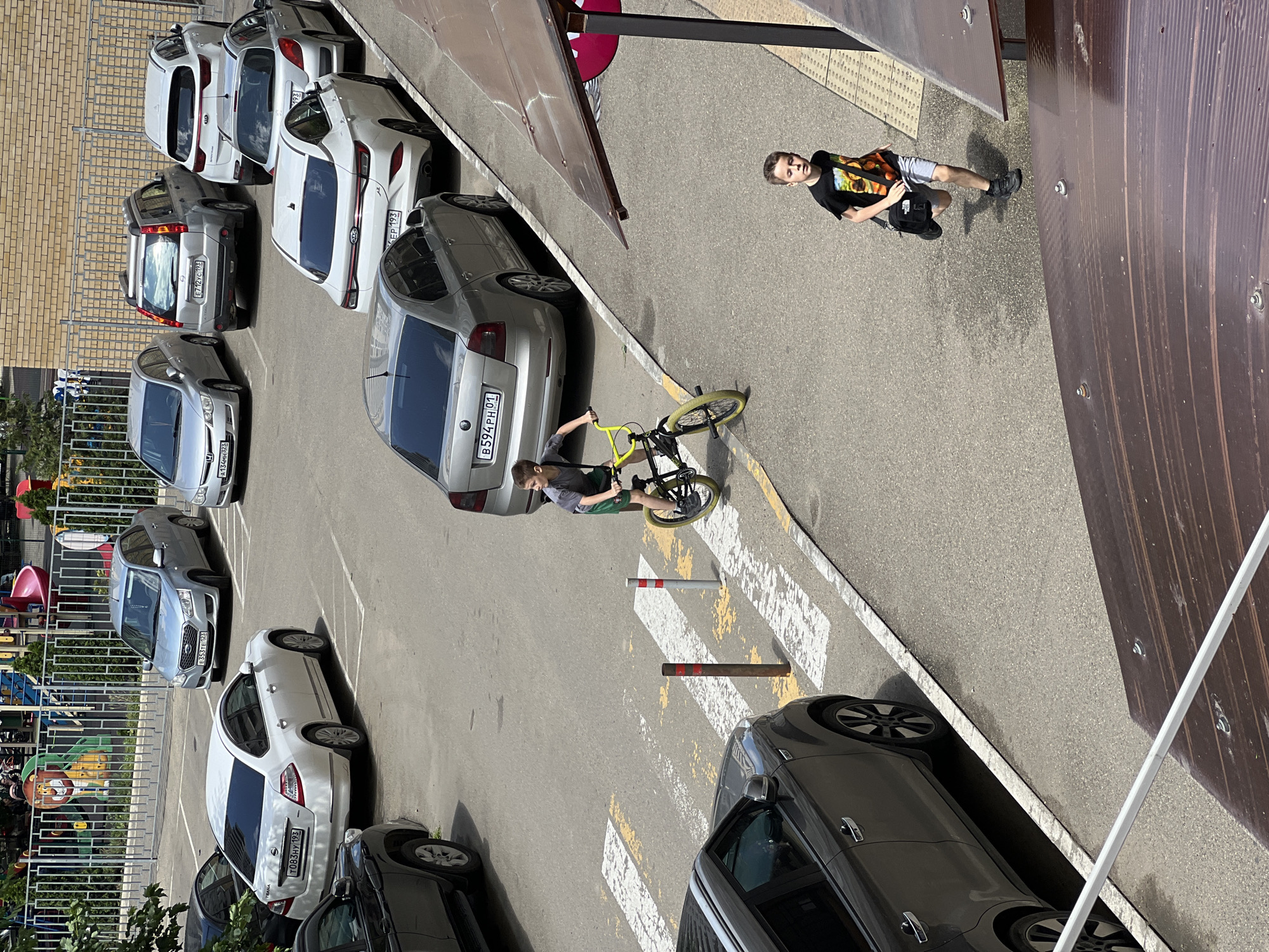 The image shows a street scene in Krasnodar, Russia. Several parked cars line the left side of the street, with a playground visible behind the cars. Two boys are present; one stands near a brown metal structure, wearing a black t-shirt, grey shorts, and black shoes, carrying a black bag. The second boy is riding a yellow bicycle on a crosswalk. A grey car with license plate B594PH01 is parked near the crosswalk. The environment is sunny, with clear visibility. The license plates on the cars suggest the location is Russia.