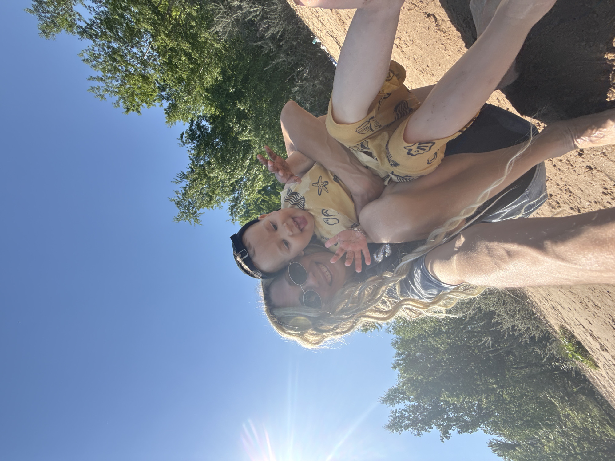 The image shows a person holding a young child outdoors on a sunny day. The person has blonde dreadlocks and is wearing sunglasses. The child is wearing a yellow outfit with a black cap. They are standing on a sandy surface, and there are trees and a clear blue sky in the background.
I do not have access to location data to confirm this is in Rusanovo, Russia.