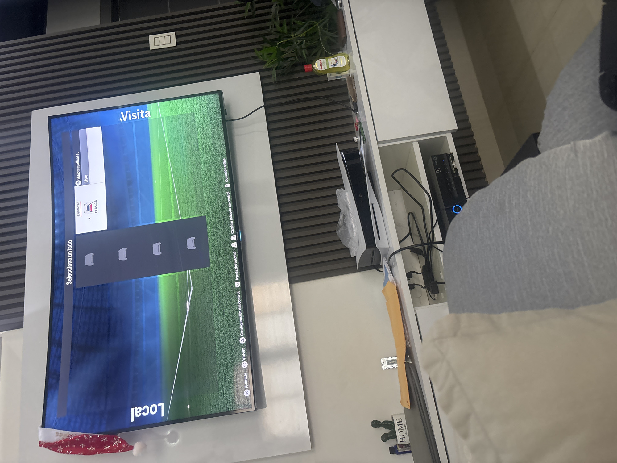 An overhead shot captures a television displaying a sports video game menu, likely a soccer game, with options for "Local" and "Visitor" teams. The screen shows a soccer field with a stadium in the background, and a menu prompting the user to "Select a side." Controller icons are visible, indicating typical game controls. A red and white Santa hat with snowflakes is perched on the top left corner of the television frame. Below the television, a white entertainment unit holds a PlayStation 5 console, a black media player, and various other items including a bottle of Huggies baby wipes, a plant, and a decorative "HOME" sign. The overall environment appears to be a living room.