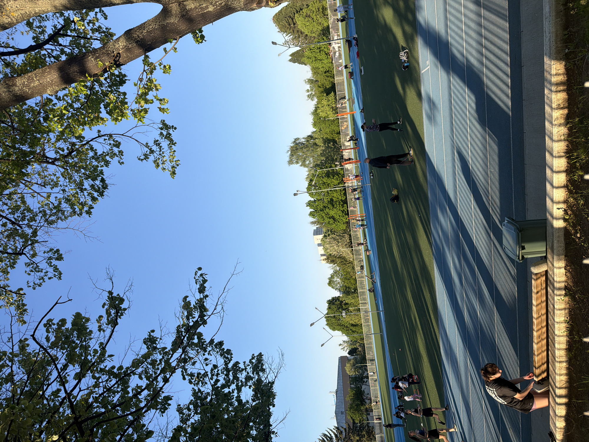The image depicts a sports complex in Sofia, Bulgaria. A blue running track is visible, with several people present, some of whom appear to be athletes warming up or stretching. There are trees framing the image, and a clear blue sky is above. There is a bench located at the right side of the image and some shadow lines are on the running track.