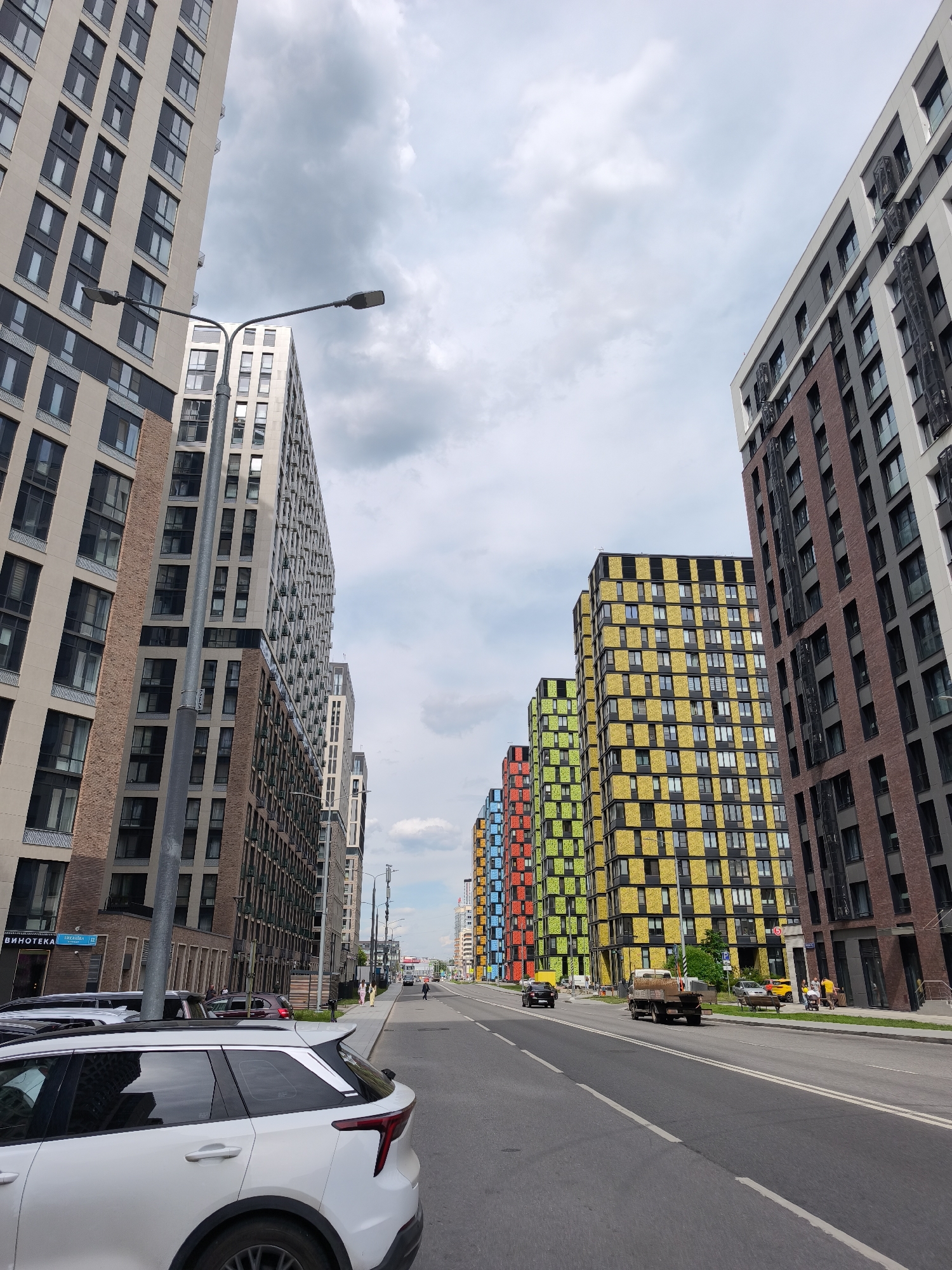 This image presents a street-level view of a modern urban landscape in Moscow, Russia, characterized by contemporary high-rise residential buildings. The scene unfolds under a partly cloudy sky, suggesting a daytime setting.
The street, an asphalt road with clear lane markings, extends into the distance, flanked by tall apartment blocks on both sides. In the immediate foreground on the left, a white SUV is parked, facing away from the viewer, with other cars parked behind it. A prominent streetlamp with two lights stands in the mid-ground on the left, extending over the road. The buildings on the left side feature facades primarily in beige, light gray, and dark gray, with some sections of brown brick, and numerous rectangular windows. A sign on one of these buildings is visible, displaying "ВИНОТЕКА" (VINOTEKA, meaning wine shop) and "ГРЕКОВА 18" (GREKOVA 18).
On the right side of the street, closer to the foreground, a tall building with a dark brown brick and dark gray facade rises. Further down the street on the right, a striking row of apartment buildings catches the eye with their colorful checkerboard patterns, alternating between bright yellow, lime green, orange, and red panels with dark gray or black sections.
The road shows typical city activity, with a truck and another car driving away from the viewer in the right lane. Pedestrians can be seen walking along the sidewalks on both sides of the street in the mid-ground, and one person is visible sitting on a bench on the right. The overall impression is that of a vibrant, active urban environment with distinct modern architectural styles.