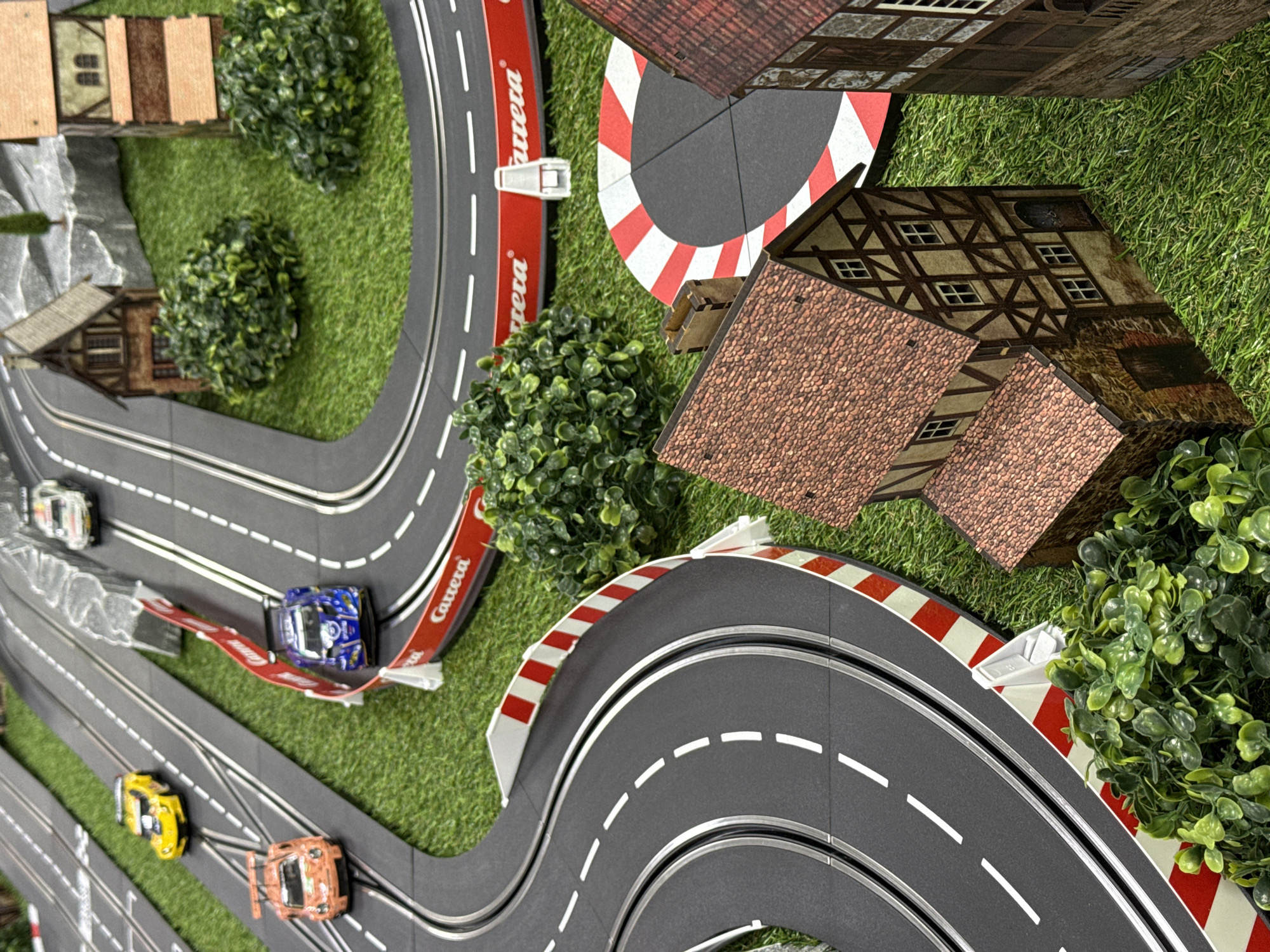 This overhead, slightly angled shot presents a detailed miniature slot car racing track setup, identified by the "Carrera" brand name visible on the red and white guard rails. The grey, multi-lane track features white dashed lines, mimicking a real race circuit.
Four slot cars are visible on the track: a white car towards the top left, a blue car on an elevated section, and yellow and orange cars further down the track. These cars appear to be sports car models, positioned as if in the middle of a race or poised to start.
The track is integrated into a miniature landscape, predominantly covered with artificial green turf simulating grass. Scattered throughout the scene are clusters of miniature green bushes and some grey rock formations, adding to the naturalistic feel. Several miniature buildings, designed in a traditional European style with timber-framed walls and red tiled roofs, are strategically placed around the track. These structures contribute to the diorama's sense of a town or countryside setting.
The scene depicts a meticulously constructed hobbyist's slot car environment. There are no people visible, and the time of day is not discernible, suggesting an indoor setup. The location context for this media file is Sirius, Russia.