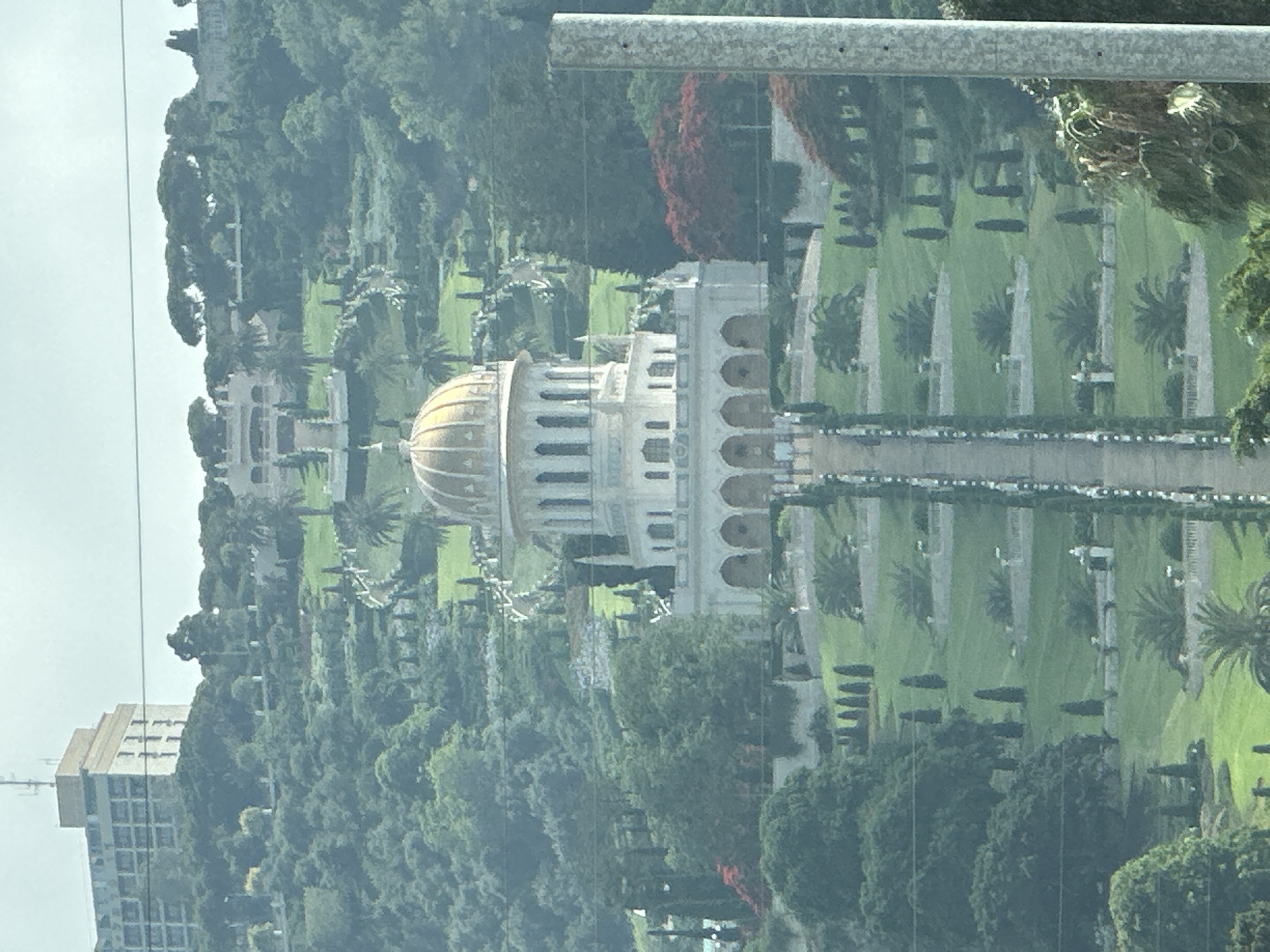 This image captures a scenic view of the Baháʼí World Centre located on Mount Carmel in Haifa, Israel. The central focus is the magnificent Shrine of the Báb, a building with a distinctive golden dome, surrounded by meticulously landscaped terraces and gardens cascading down the mountainside. The architecture is elegant, characterized by white stone structures, arched walkways, and numerous symmetrically placed cypress trees and other lush greenery. In the background, a modern multi-story building with antennas on its roof can be seen on the upper slopes of the mountain. The overall scene depicts a harmonious blend of religious architecture, natural beauty, and urban development on the slopes of Mount Carmel.