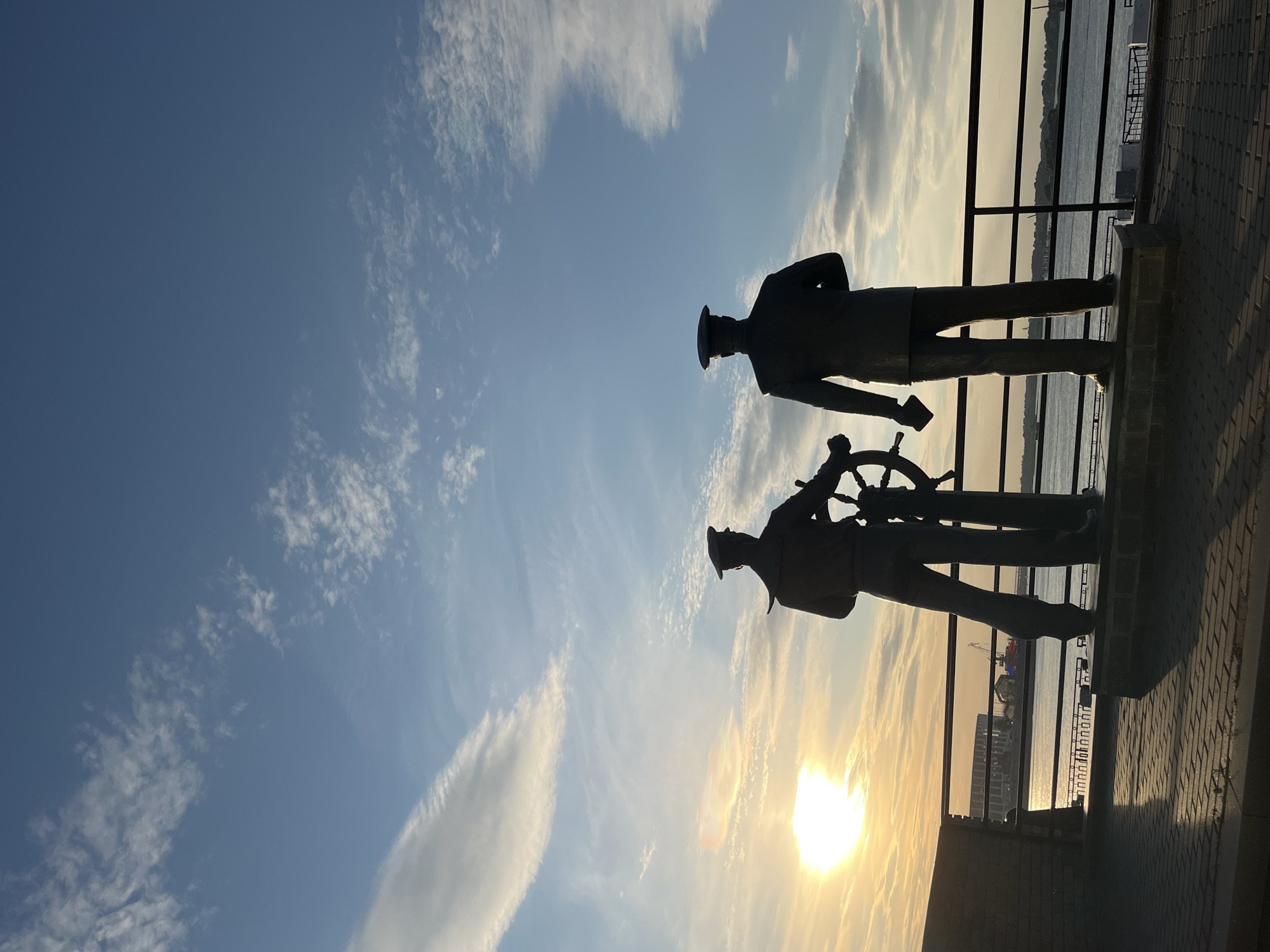 The photograph depicts two silhouetted statues of men in naval uniforms standing on a paved promenade overlooking a body of water. The statue on the left is positioned at a ship's helm, while the statue on the right stands beside it, looking out towards the water. The sun is setting on the left side of the frame, casting a warm glow and long shadows across the promenade. In the background, across the water, are industrial buildings and a crane. The sky is a gradient of blue with scattered white clouds. This scene is located on the Volga River embankment in Nizhny Novgorod, Russia, likely near the confluence of the Volga and Oka rivers.