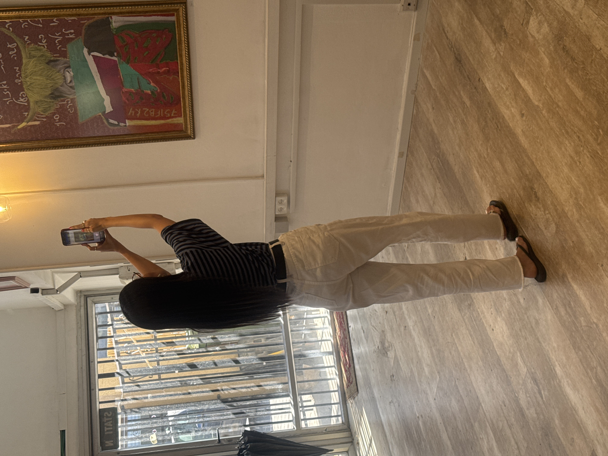 A woman in a striped shirt and white pants is taking a photograph with a smartphone in what appears to be an apartment in Tel-Aviv, Israel. The woman has long, dark hair and is wearing flip-flops. In the background, there's a painting hanging on the wall. A window is visible, with bars, suggesting a view of Tel-Aviv, and an umbrella is resting in front of the window. The floor is a light, wood-like material.