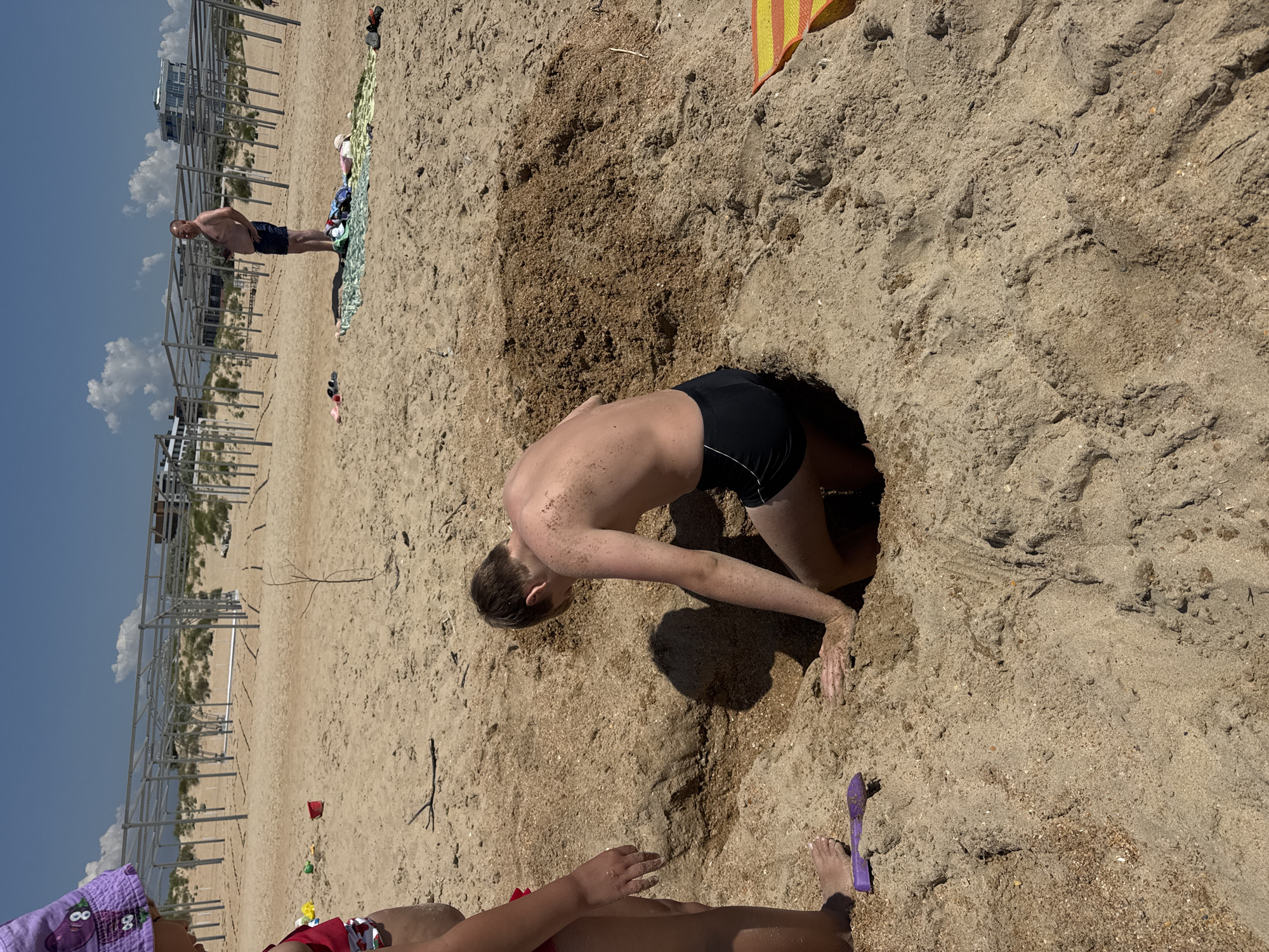 This is a photograph taken on a sandy beach, likely in Vityazevo, Russia, given the context. The image features several people. In the foreground, a child wearing a purple hat with cartoon characters and a red swimsuit is partially visible, with their hands resting on the sand. To the left, another person, possibly an adult, is seen from the waist down, also in swimwear.
The central focus of the image is a young person, dressed in black swim trunks, who is energetically digging into a large hole in the sand. Their body is contorted as they reach into the hole, appearing to be engrossed in the activity. The sand is a light brown color, with areas of darker, damp sand where the digging is taking place. A bright yellow and red striped towel is visible in the upper right corner of the frame, suggesting a relaxed beach day.
In the background, a metal structure, possibly part of a beachside attraction or construction, stands against a clear blue sky with scattered white clouds. The overall impression is one of a sunny day at the beach, with people enjoying recreational activities. The composition and lighting suggest the photo was taken during daylight hours, likely mid-day. No visible text is present in the image.
