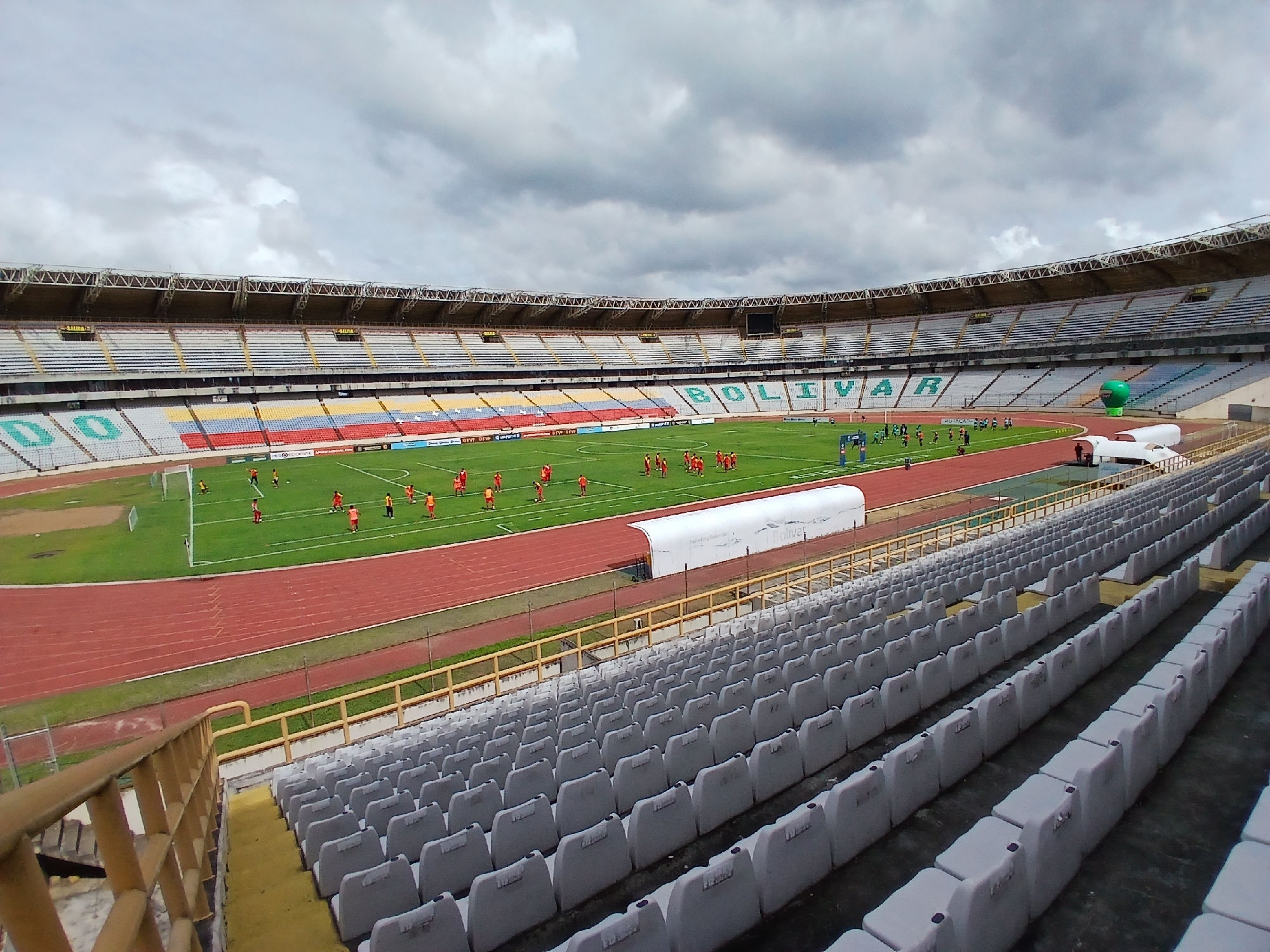 The image shows an elevated view of Estadio Cachamay in Ciudad Guayana, Venezuela. A football (soccer) match or practice session is underway, with players in red and blue uniforms on the green field. A red running track surrounds the field. A large, partially inflated green balloon-like object is visible near the stadium's stands. The stands have multiple tiers of seating, with the colors of the Venezuelan flag (yellow, blue, red) displayed on one section. The word "BOLIVAR" is spelled out across a section of seating. The sky is overcast. The foreground features rows of empty gray stadium seats.