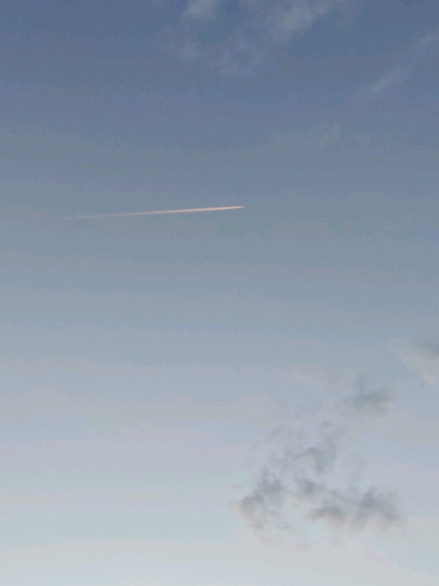 The image depicts a clear sky with a faint contrail left by an aircraft, stretching horizontally across the upper portion of the frame. The sky transitions from a light blue at the top to a paler, almost white hue towards the bottom. Scattered clouds are visible, concentrated in the lower right quadrant of the image, appearing wispy and greyish. The overall lighting suggests either early morning or late afternoon. The specific location in Rubtsovsk, Russia, is not determinable from the visual information presented. There are no people, specific objects, or actions identifiable within the scene.