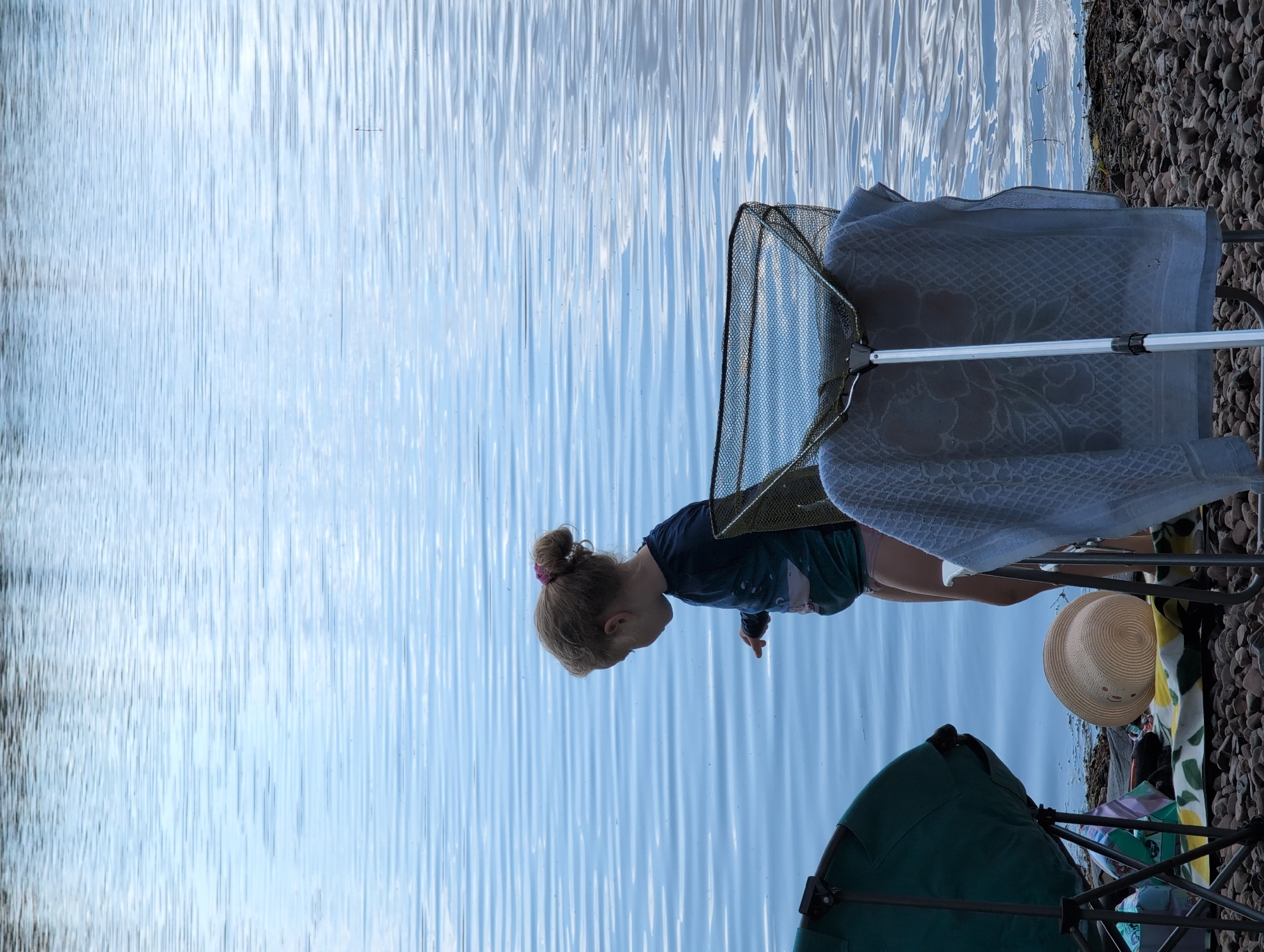 In an outdoor setting next to a body of water, a young girl with her hair in a bun is fishing with a net. She is wearing a dark t-shirt and green shorts. A fishing net is held up in front of her. Beside her are a straw hat and a folded blanket. A fishing chair and a green tent are also visible, suggesting a recreational fishing trip. The water is calm and reflects the sky, indicating fair weather. The scene appears to be during daylight hours, possibly late afternoon, given the lighting. The location is identified as Petershagen, Germany.