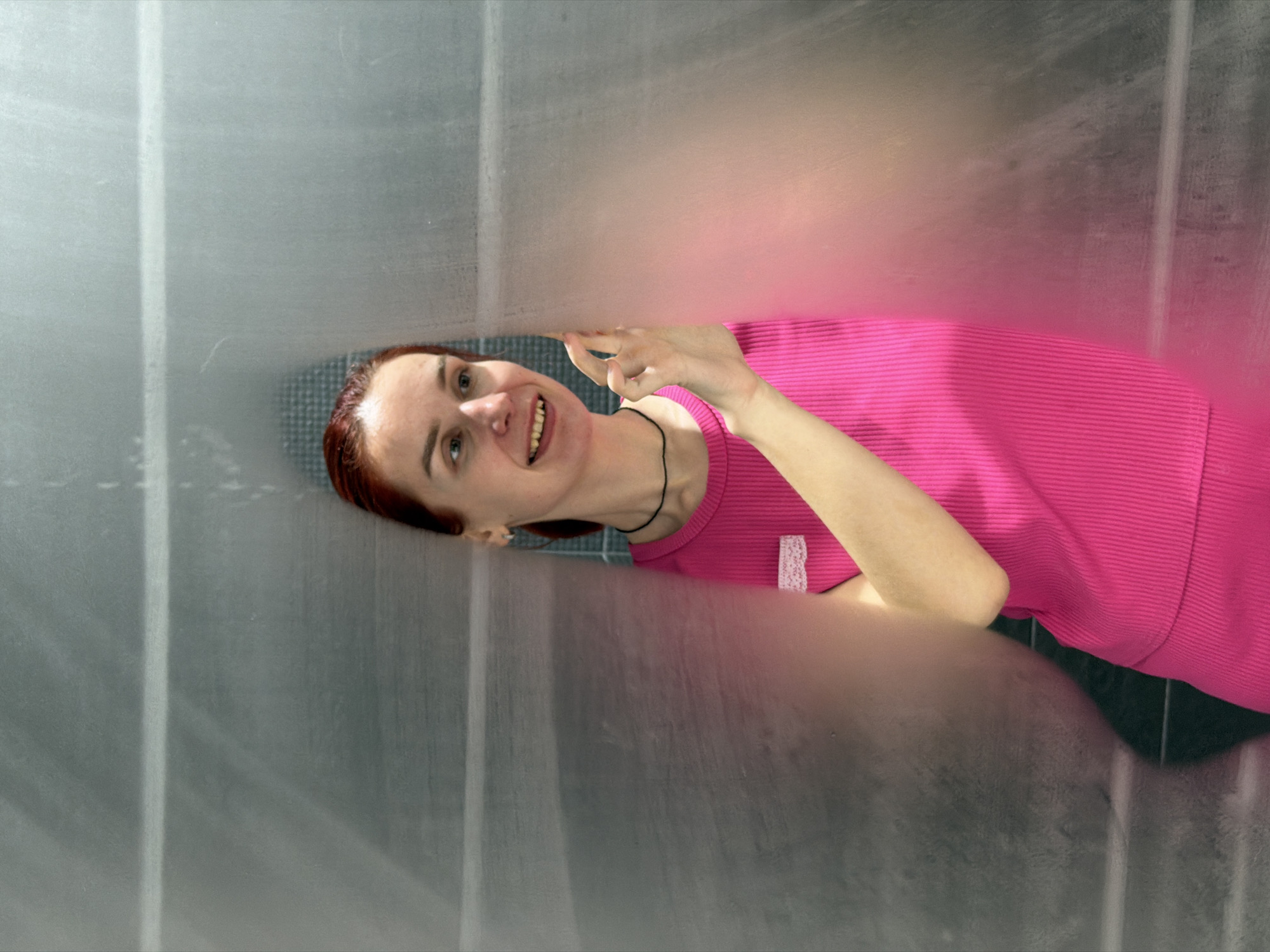 A woman with red hair and a pink shirt is smiling and looking up through a gap in a gray, textured surface. Her hand is visible near the gap. The location is likely Chaikovsky, Russia, based on potential metadata not visible in the image.
