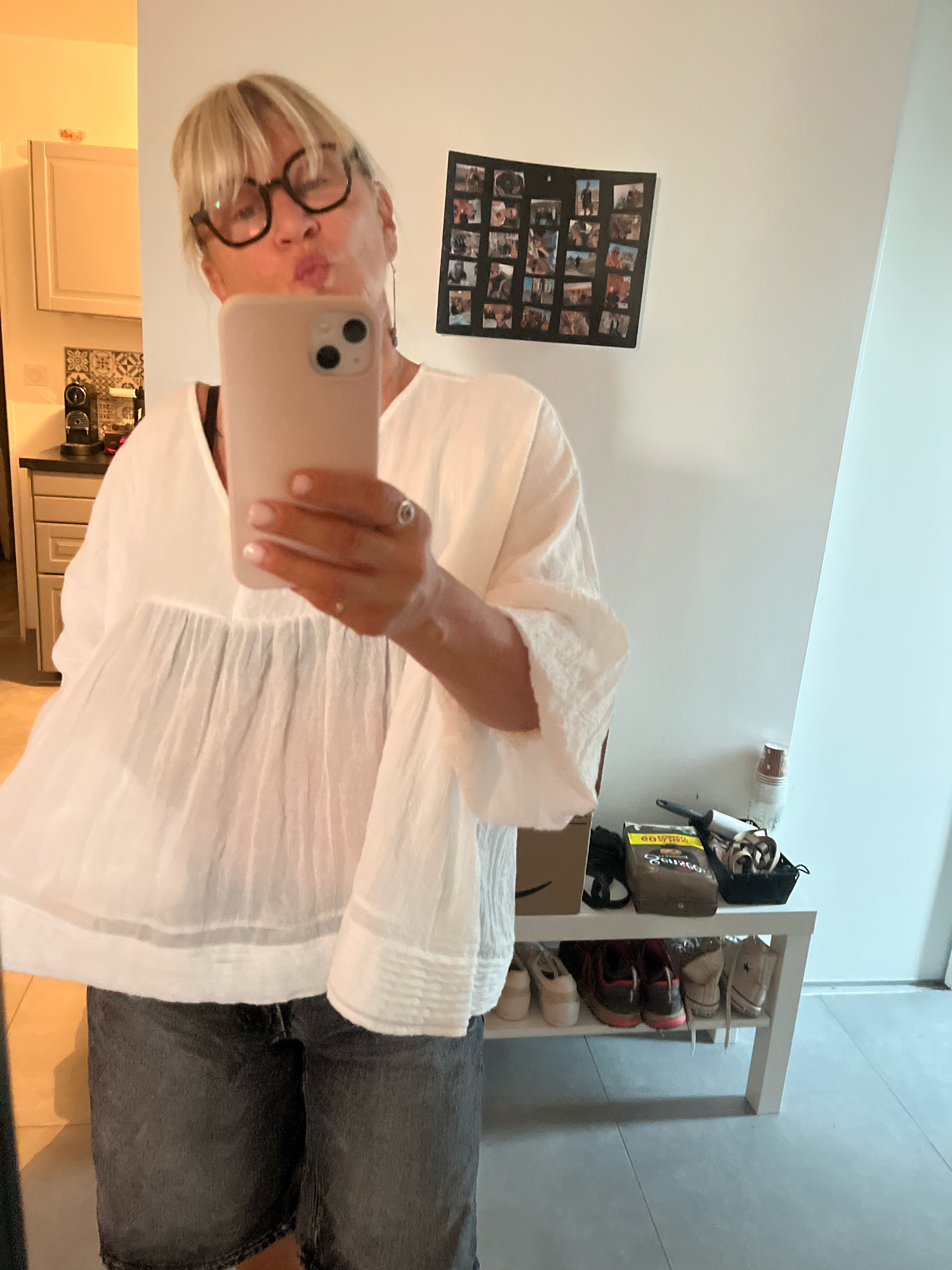 The image shows a medium-sized woman with blonde hair and glasses taking a selfie in a well-lit room. She is wearing a white peasant blouse and denim shorts. In the background, there are kitchen cabinets, a collage of photos on the wall, and a shelf with shoes and other items. The scene appears to be a domestic interior, possibly a home. There is no indication that this image is related to Chambœuf, France.