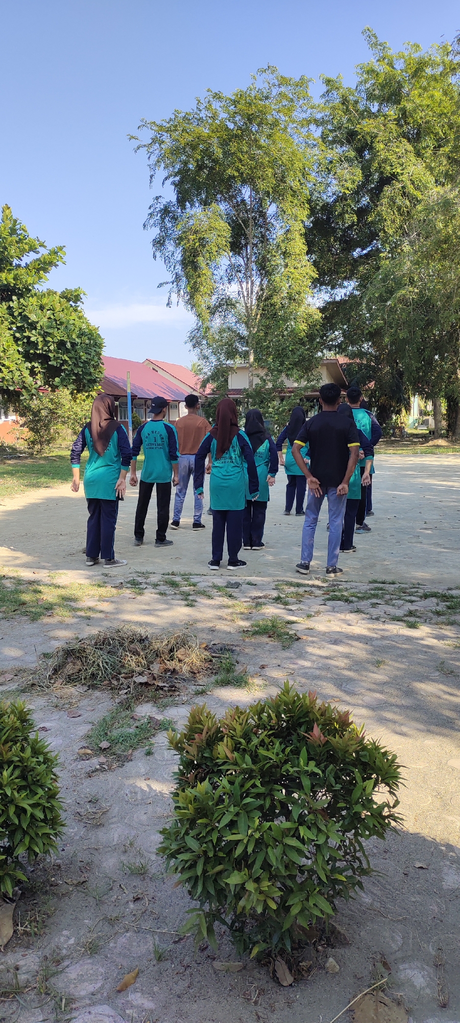 A group of individuals, some wearing teal shirts with black sleeves and dark pants, and others in casual wear, are gathered outdoors. The individuals in teal shirts appear to be students, with two wearing hijabs. One person in a black t-shirt and blue jeans stands facing away from the camera. They are on a sandy or gravel surface, with some dried grass and leaves scattered around. In the background, a red-roofed building with white walls is visible, surrounded by trees. The scene appears to be a schoolyard or outdoor recreational area, potentially in Langnibong, Indonesia, given the architectural style and vegetation. The sky is clear and blue.