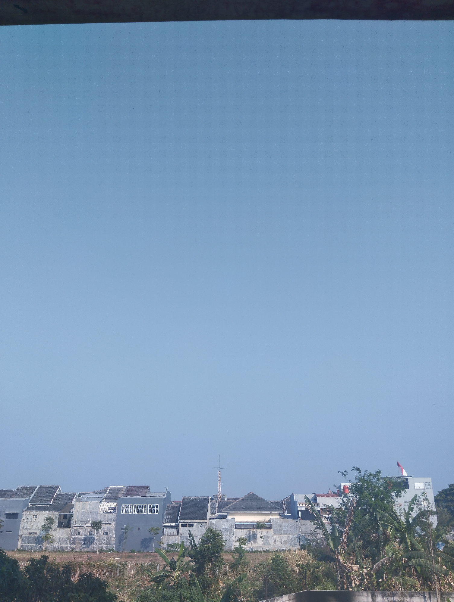 The image captures a panoramic view of a residential area in Madiun, Indonesia, under a clear, pale blue sky. In the foreground, a dark, horizontal element, possibly a windowsill or ledge, partially obscures the top of the frame. Below this, a line of houses forms the main subject. These houses are of varying designs and sizes, constructed with gray walls and dark tiled roofs. Some have multiple stories, while others are single-level. Windows are visible on many of the structures, and a few appear to have clothes drying on balconies. A flagpole with an Indonesian flag is visible on the roof of one of the buildings on the right side of the frame.
The houses are situated behind a stretch of dry, brown grass and low-lying vegetation. Beyond the houses, a few trees with lush green foliage dot the landscape, with some taller trees on the right side partially obscuring the view of the houses. The overall impression is that of a typical suburban or semi-urban neighborhood in Indonesia, likely during the dry season given the appearance of the ground cover. The scene is peaceful and devoid of human activity, presenting a still, architectural and environmental snapshot of the location.