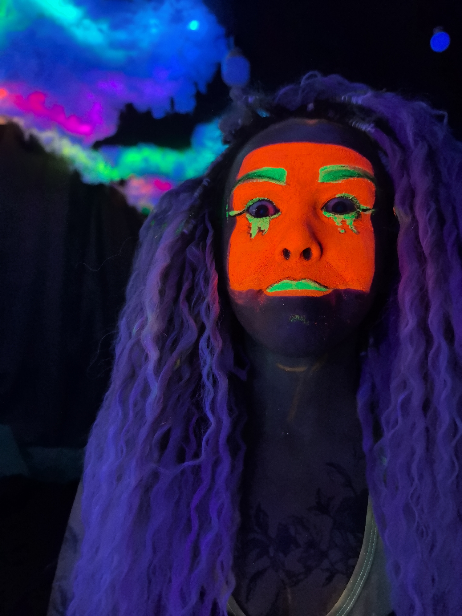 A medium shot shows a person with orange and green neon face paint and long, wavy purple hair. The face paint covers the forehead, nose, cheeks, and chin, with green accents on the eyebrows, under the eyes as if tears, and on the lips. The skin around the face paint appears dark, and a floral design is visible on the chest, suggesting a tattoo. The hair cascades down on both sides of the face. In the background, there are neon lights in shades of pink, green, and blue, suggesting a themed or decorated space. The lighting gives a surreal and artistic quality to the image. The image does not appear to be taken in Noviy, Russia. There is no evidence to suggest it was.