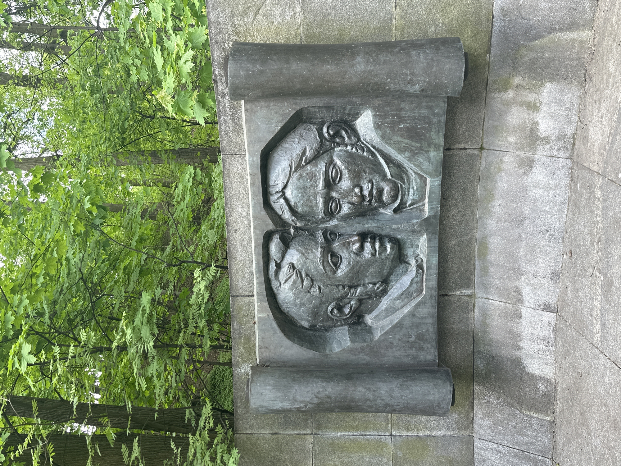 A bronze sculpture of two faces, one above the other, is set in a stone wall near a park. The faces appear to be of classical style. The sculpture is in the form of an open scroll. The background shows greenery, suggesting a park or garden setting. The location is in the city of Moscow, Russia.