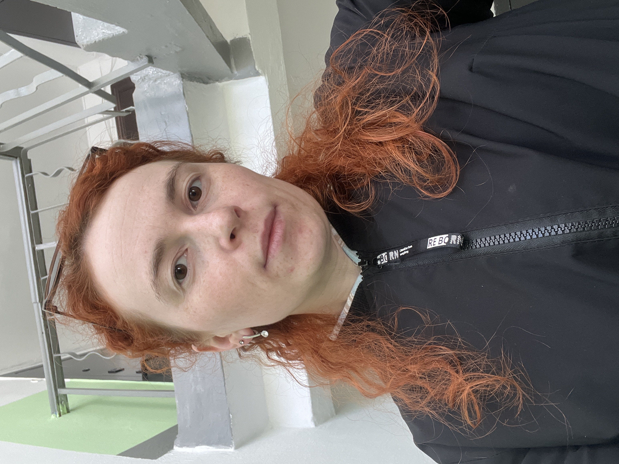 A fair-skinned person with red hair is wearing a black jacket. The jacket zipper is visible at the collar. The person is wearing glasses and a stud earring. The background shows a structure with gray and white surfaces and green accents. The location is Chaikovsky, Russia.