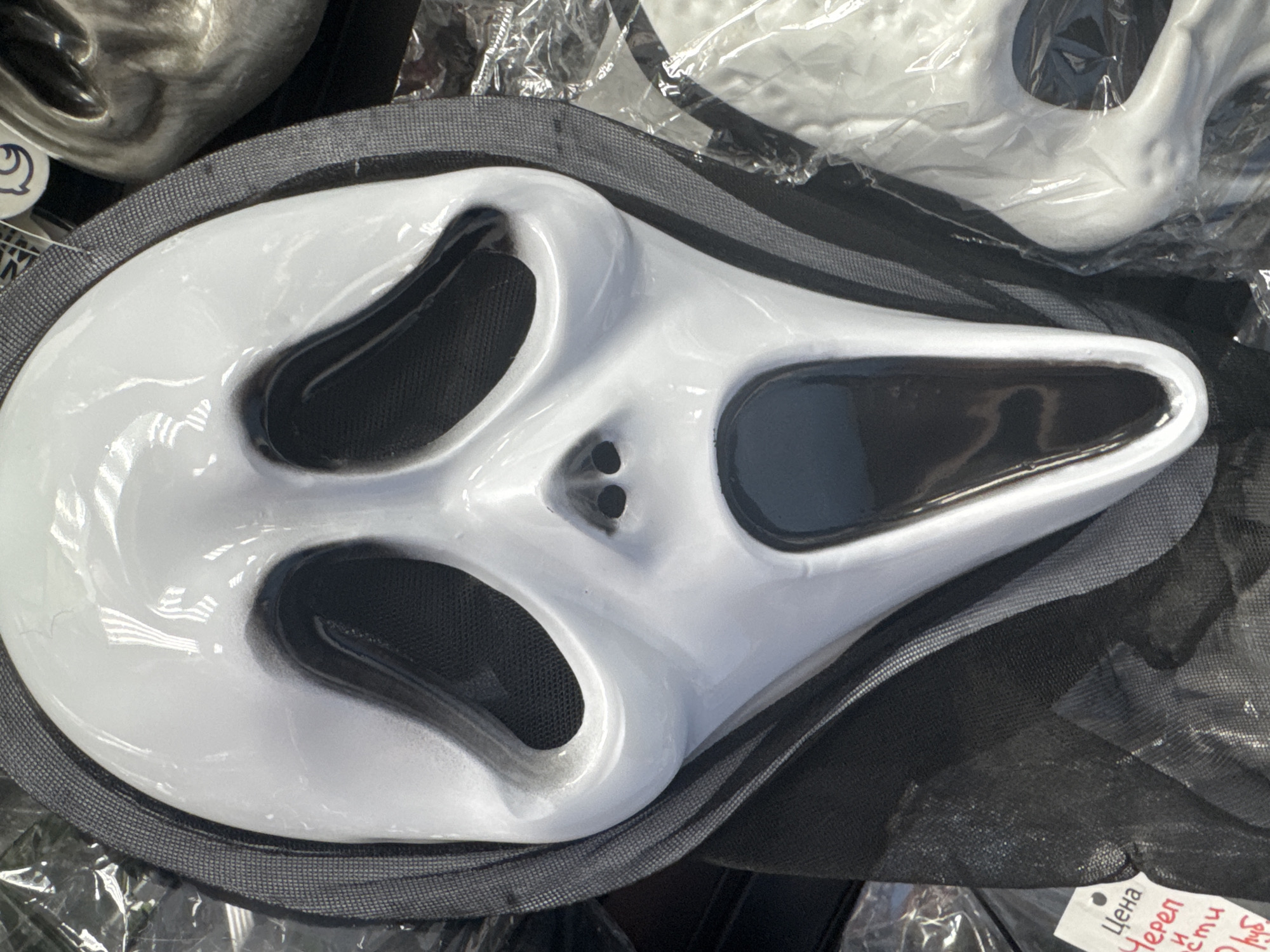 A white "Scream" mask is the main subject, lying on a gray surface with black netting underneath. The mask features prominent black eye and nose holes. A price tag is visible on the lower right. The scene appears to be indoors, possibly a store or market, based on the presence of the price tag and other items in the background. There's no direct evidence to confirm the location is Saint Petersburg, Russia, but that may be indicated by the writing on the price tag.