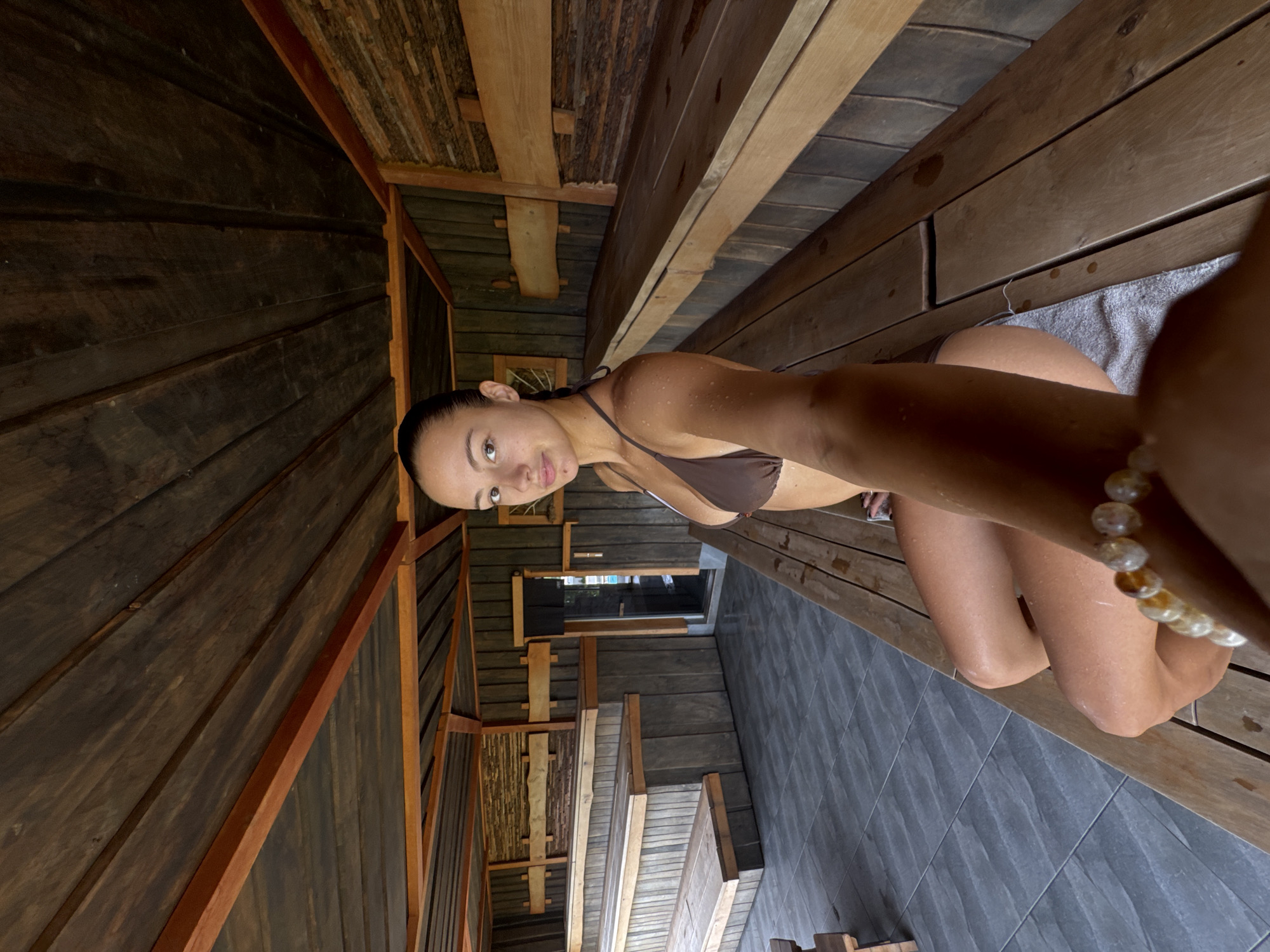 A woman is sitting in what appears to be a sauna or rustic wooden room in Kathu, Thailand. The walls and ceiling are constructed of rough-hewn wooden planks, with varying textures and finishes. The woman is wearing a brown bikini and a beaded bracelet. She appears to be holding a camera or phone out to take a selfie. The background shows tiled flooring.