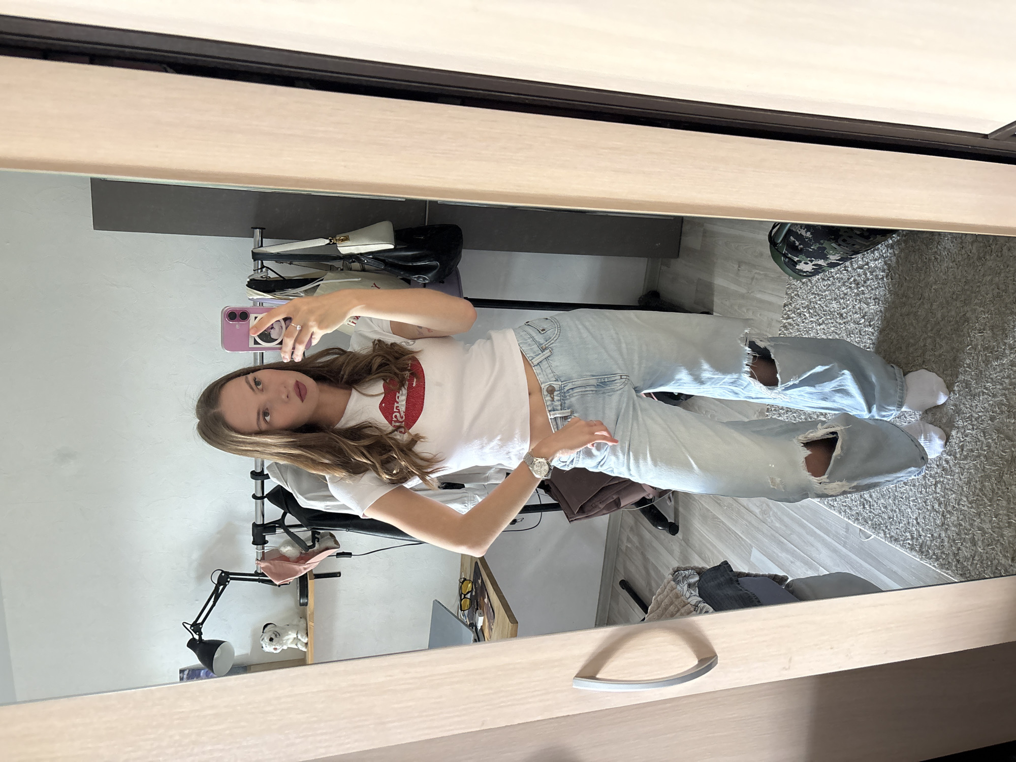 A young woman with long, wavy brown hair stands in front of a mirror, taking a selfie. She is wearing a white t-shirt with a red lip graphic and light-wash distressed jeans. Her left arm is raised, holding a purple smartphone. She is also wearing a silver watch on her left wrist.
The setting appears to be a bedroom or dressing area. In the reflection of the mirror, we see a clothing rack with some items of clothing, a black desk lamp, and a small white dog figurine on a shelf. There is also a floor rug and a camouflage-patterned backpack visible.
The lighting suggests it is daytime. The woman's expression is neutral as she looks into the camera. The overall mood is casual and relaxed. The location is specified as Perm, Russia.