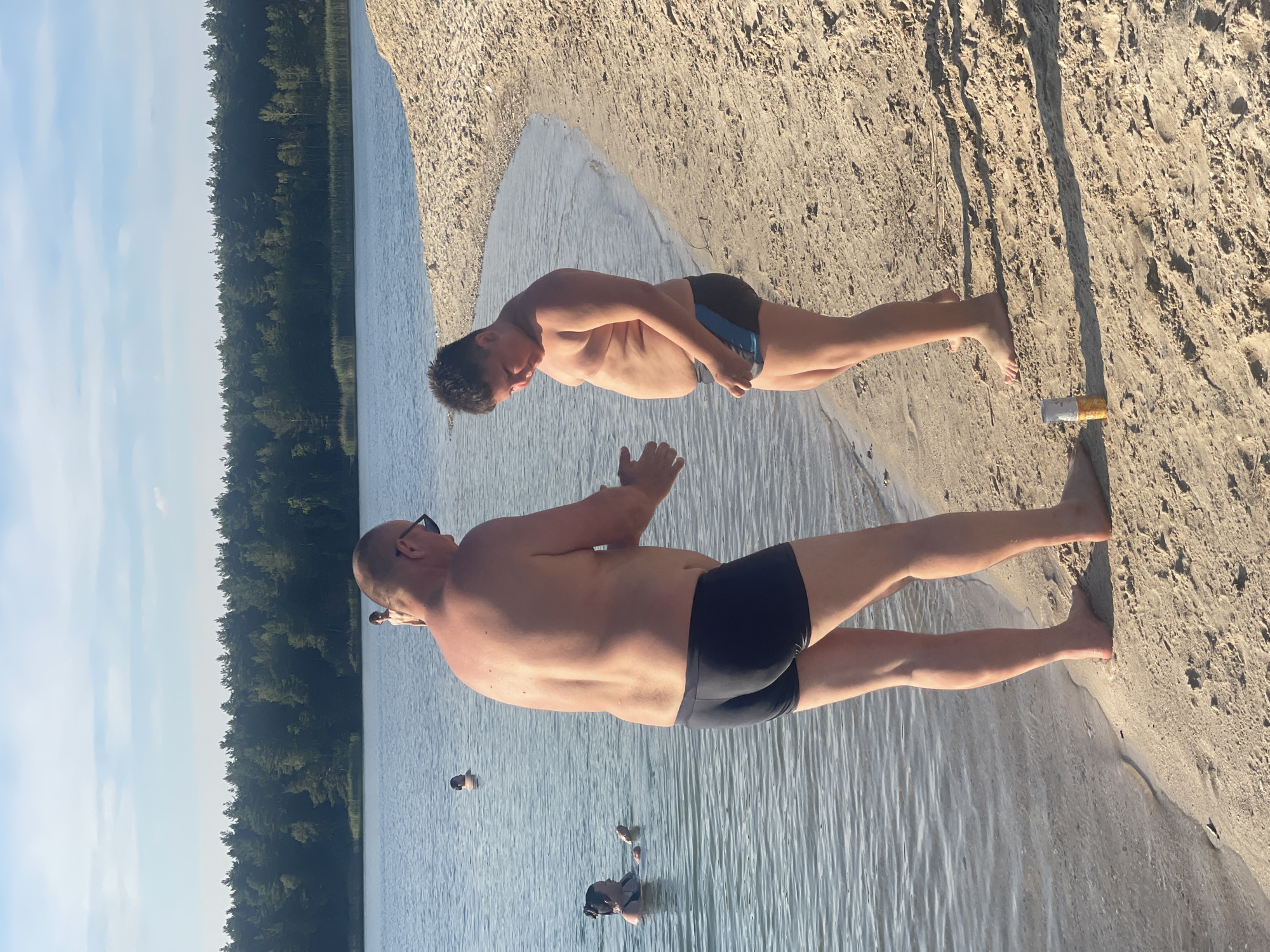 The image shows two men in swim trunks at the edge of a body of water, with a sandy beach and a forest in the background. The image is oriented vertically. The man in the foreground, closer to the viewer, is facing away and looking out at the water. He has sunglasses on and appears to be an adult. The second man is further back, facing towards the viewer and looking down. He is thinner than the first man and appears to be younger. The water is calm and reflects the sky, which is a pale blue with some white clouds, suggesting it is daytime, likely morning or afternoon. The sun casts shadows on the sand, indicating a sunny day. There are a few other people in the water in the distance, partially submerged. On the sand near the foreground man's feet, there is a discarded cigarette butt. The overall scene suggests a relaxed day at the beach or lake.