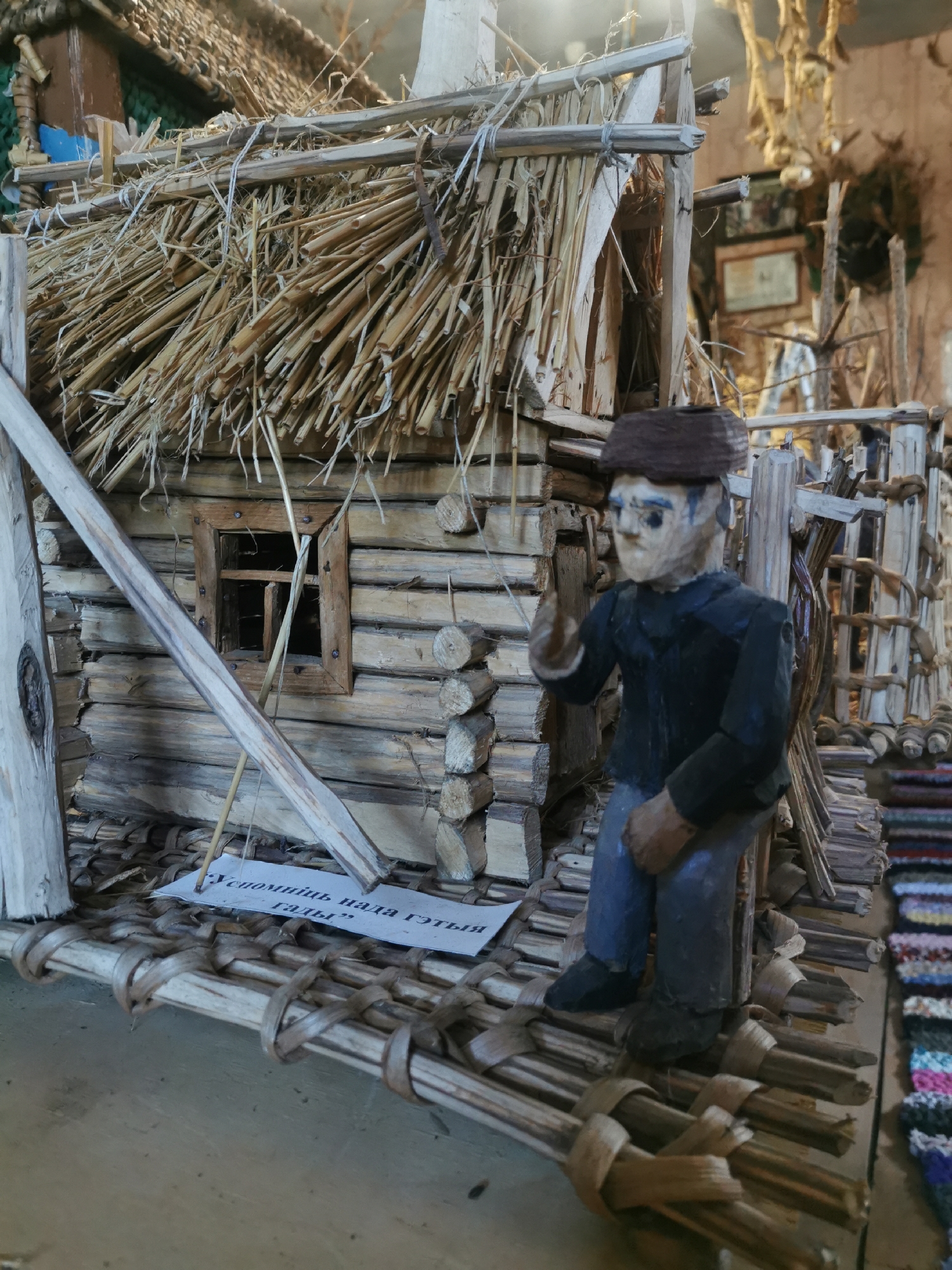 An outdoor, medium shot captures a meticulously crafted diorama of a traditional Belarusian dwelling, likely in the village of Шчарчоўскі сельскі Савет, Belarus. The central focus is a miniature log cabin with a thatched roof, constructed from small wooden logs and straw. A small, recessed window with a wooden frame is visible on the cabin's facade. In front of the cabin, a small, carved wooden figure of a man stands on a wooden platform, gesturing with his right hand. The figure wears a dark jacket and blue trousers, with a dark hat. A piece of paper with Cyrillic text is placed on the platform near the figure. The diorama is set against a backdrop that suggests an indoor display area, with other rustic artifacts and woven textiles visible in the background. The overall impression is one of historical preservation and folk art.