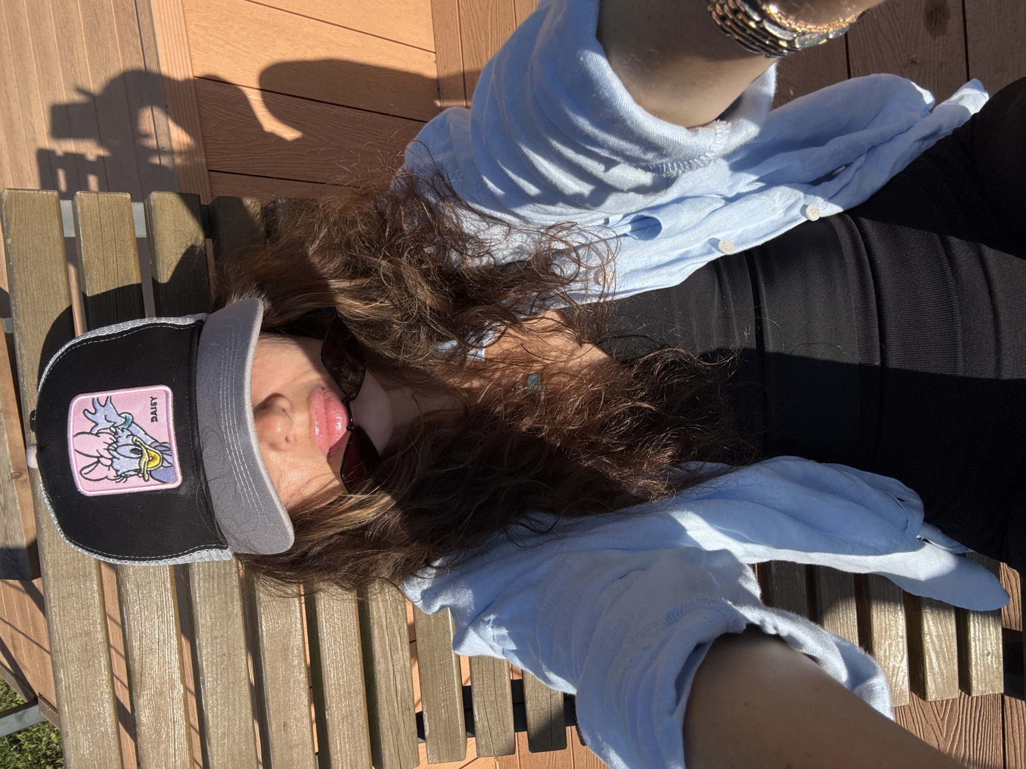 A person wearing a light blue button-up shirt over a black top and a trucker hat with a Daisy Duck patch is sitting on a wooden bench. Sunglasses are perched on their mouth. The person's long, dark, curly hair is spread out. They are outdoors, with wooden decking and planks visible in the background. The scene is bathed in sunlight, casting shadows. There is no indication that this image was taken in Moscow, Russia.