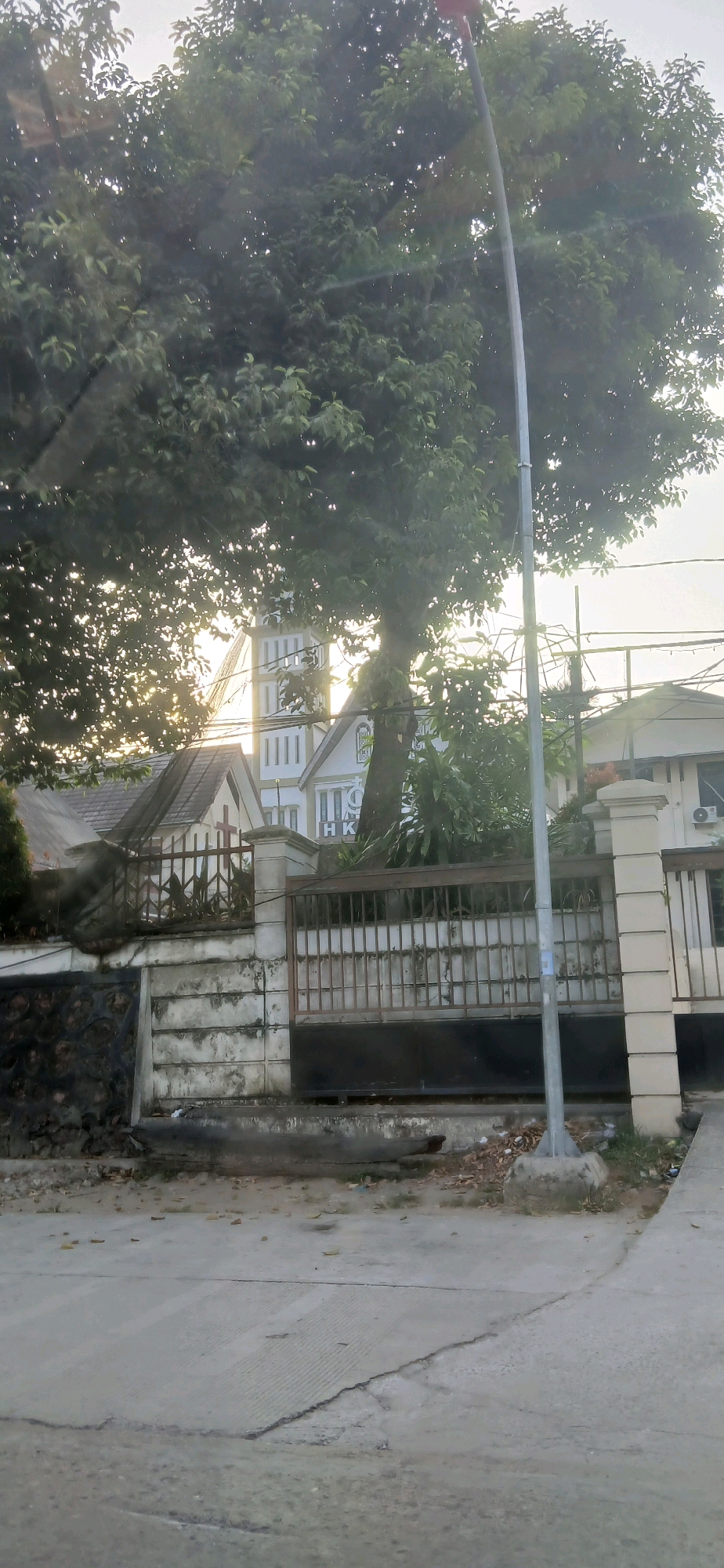 The image is a medium-shot photo of a street scene in Samarinda, Indonesia, taken during the day. A large tree with dense green foliage dominates the left side of the frame, with sunlight filtering through its leaves. Behind the tree, the top of a white and green building with a tall tower is visible. The building appears to be a church, indicated by a red cross visible on a smaller adjacent structure to the left. A tall, gray lamppost stands in the foreground, slightly off-center to the right, with its base on a concrete platform. In front of the building, a metal fence with vertical bars and a black gate separates the property from the street. The street in the foreground is paved with concrete, showing some cracks and wear. The overall impression is of a residential or institutional area.