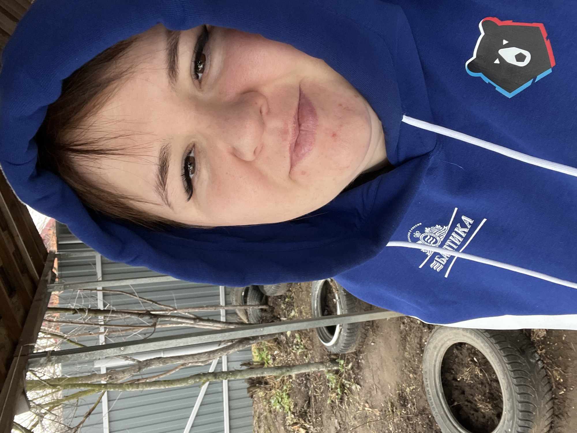 A woman wearing a blue hoodie with a bear logo and the word "Балтика" is the primary subject. The woman is tilted slightly, and her face is visible beneath the hood. The background consists of a metal structure, bare tree branches, and several tires. The location is determined to be in Chaikovsky, Russia due to the inscription on the hoodie.