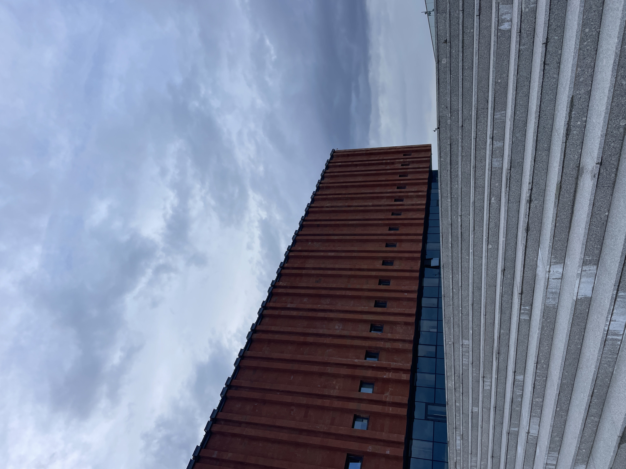 The image captures a low-angle view of modern architectural structures set against a cloudy sky. On the left, a tall, reddish-brown building dominates, characterized by prominent horizontal bands that run its full height. Small, rectangular windows are interspersed seemingly randomly along these bands. The left edge of this building appears to have dark, stepped ledges, while its right side features a sleek, dark glass facade reflecting the sky. To the right, another structure, possibly an adjacent building or a distinct part of the same complex, presents a light grey, textured surface. This surface is highly detailed with numerous parallel horizontal ridges or stripes, giving it a corrugated appearance. The sky above is overcast, filled with various shades of grey and white clouds, indicating a cloudy or partly cloudy day. No sun is directly visible. There are no people or specific activities depicted. The architectural style suggests an urban or modern city environment. No discernible text or explicit location cues are visible. The overall composition uses strong diagonal lines from the buildings contrasting with the expansive sky.