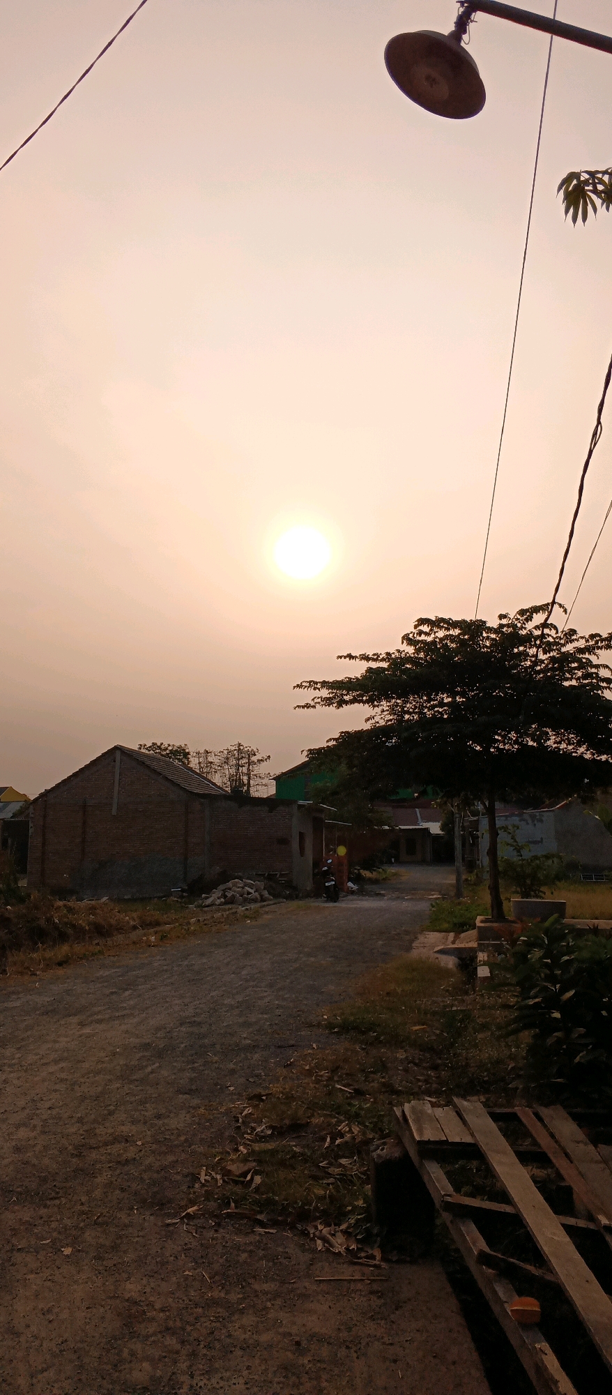 The image depicts a dusty road in the village of Watubelah, Indonesia, during a sunset. The sky is a hazy orange, with the sun appearing as a bright white orb slightly to the left of the frame's center. On the left side of the road, a partially constructed brick building stands, with some rubble and debris in front of it. Further down the road, a few more houses are visible, some with green roofs. A mature tree with dense foliage stands on the right side of the road, casting a shadow. A street lamp hangs from a pole on the right side of the image, and power lines run across the sky. In the middle ground, a motorcycle is parked near one of the houses. The road itself is unpaved and appears to be made of dirt and gravel. Some dry grass and fallen leaves are scattered along the sides of the road and in the foreground. In the lower right corner, a stack of wooden planks is visible. The overall impression is one of a rural village at dusk.