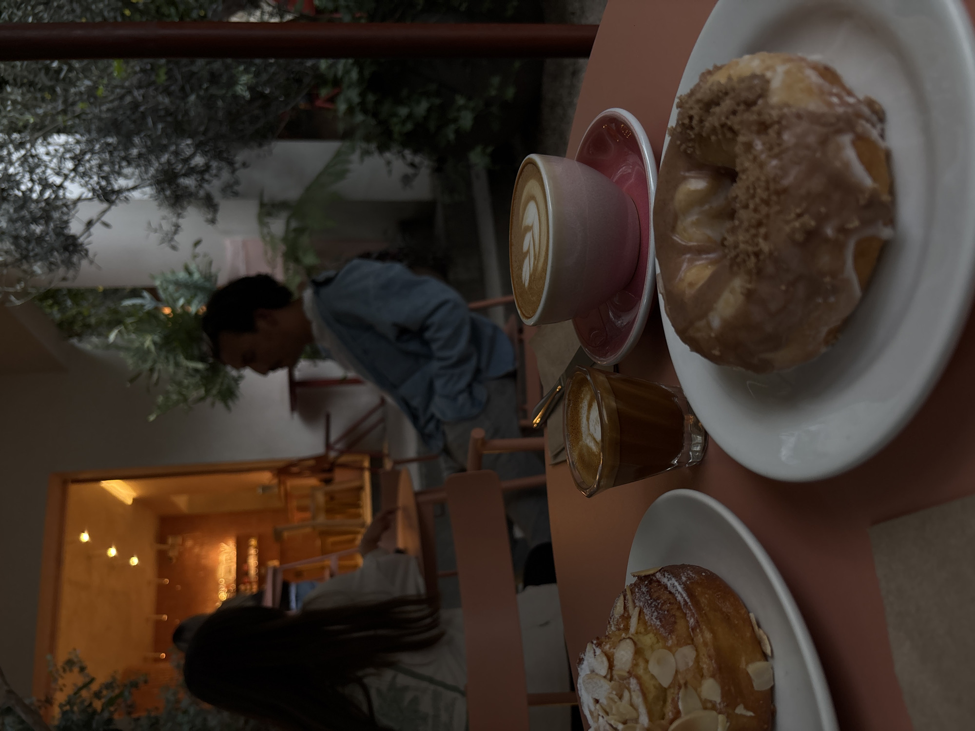 The image presents a table with several items in the city of Unknown, Unknown. A cup of latte art sits on a pink saucer next to a glazed donut with sprinkled crumbs on a white plate. A small glass of coffee is beside them. Below these, another white plate holds an almond croissant. In the background, a blurred person in a blue shirt stands in a light-filled room with several chairs visible. The outside area shows foliage.