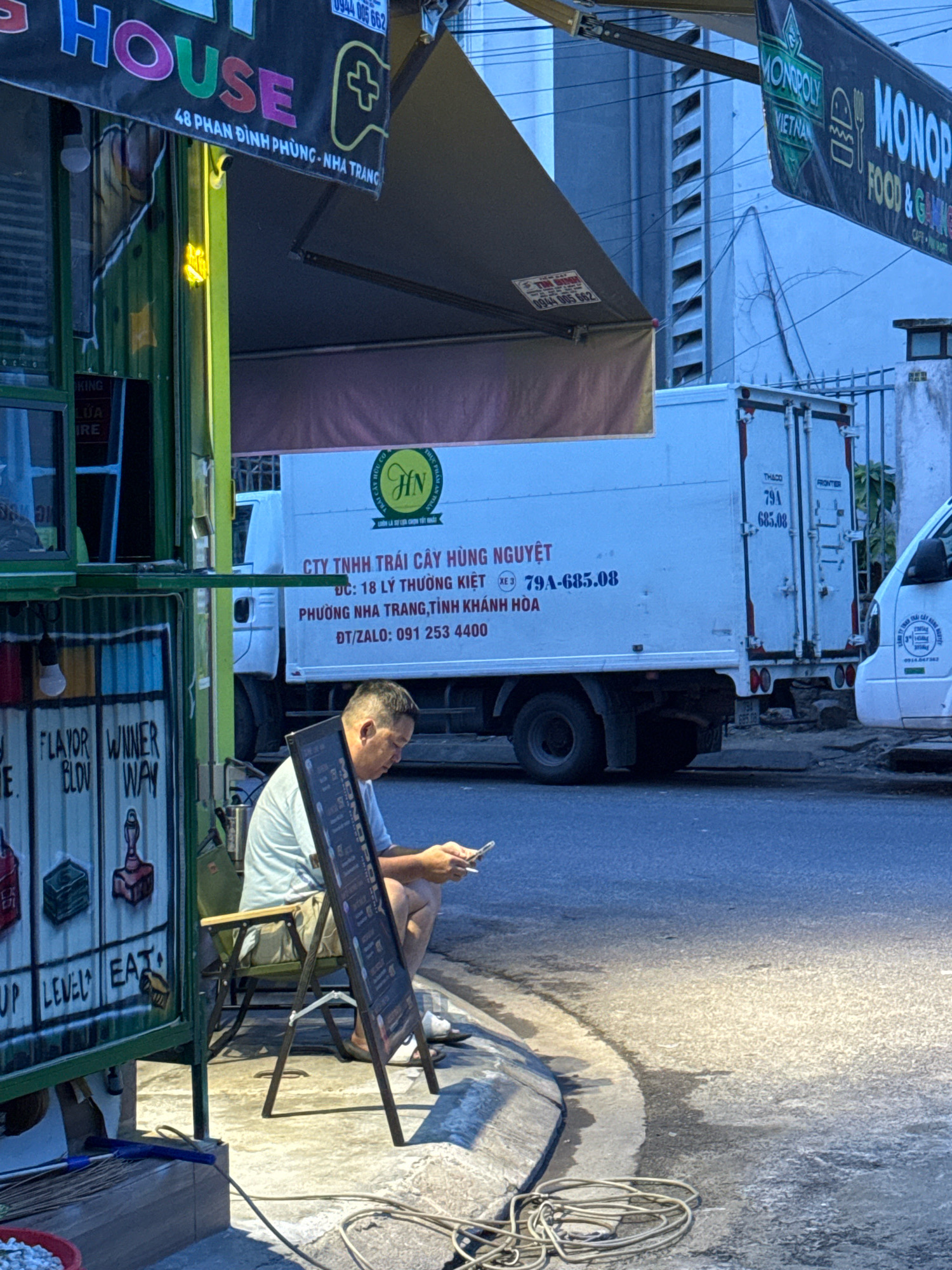 An outdoor scene in Nha Trang, Vietnam, shows a man sitting on a folding chair and looking at his mobile phone. He is positioned near a green stall with colorful paintings of objects like slot machine symbols and trophies. A black easel sign with text is placed in front of him. In the background, a white refrigerated truck is parked, displaying text in Vietnamese related to a fruit company, including a phone number and address on Phuong Nha Trang street. Other elements in the scene include awnings, power lines, and a portion of another white vehicle.