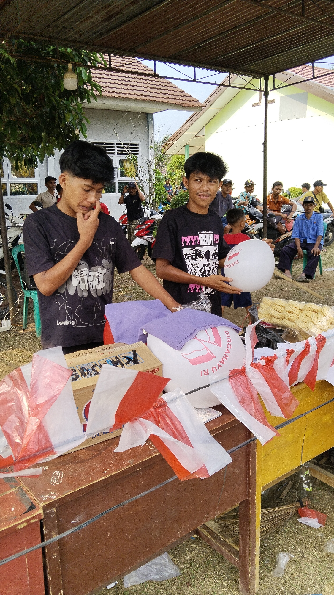 Two young males, one with dark hair styled upwards and wearing a black t-shirt with a graphic design of cartoon-like figures, is looking down with his hand on his chin, appearing to be in thought. The other young male, also with dark hair styled upwards and wearing a black t-shirt with a graphic of faces, is holding a white balloon. They are standing behind a wooden table in what appears to be an outdoor setting, possibly in Margatunggal, Indonesia, indicated by the presence of several people in the background on motorcycles and buildings with tiled roofs. The table is covered with various items including boxes, purple cloth, and red and white Indonesian flags draped over them. In the background, more people are visible, some on motorcycles, and buildings with open windows and tiled roofs are present. The ground is covered with dry grass. The overall scene suggests a community gathering or an outdoor market.