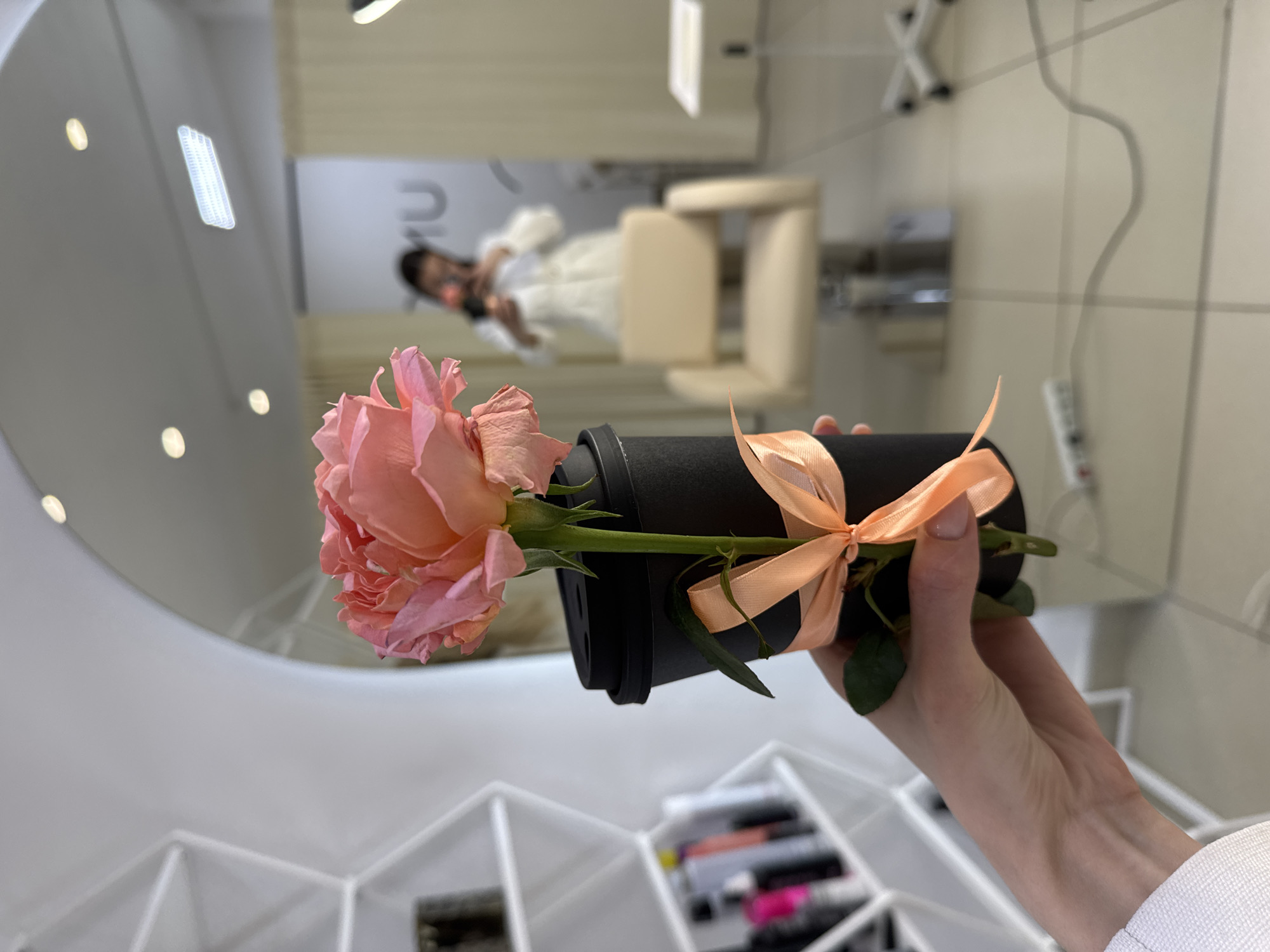 A person is holding a pink rose wrapped in a black container with a peach-colored ribbon in what appears to be a makeup studio. The reflection in the mirror shows another person wearing a white dress, seated in a beige salon chair. The environment includes makeup organizers and salon equipment, suggesting the location is most likely a beauty salon. Based on the information provided, it is located in Chaikovsky, Russia.