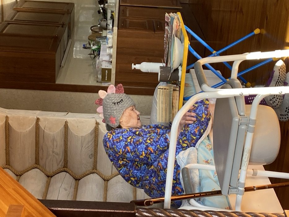 In a residential interior within the city of Кукуи, Russia, an elderly female individual is depicted seated, leaning forward with her head bowed, suggesting a state of rest or sleep. She wears a blue robe with a floral pattern and a grey knitted hat featuring pink protrusions, embroidered closed eyes, and a pink star. She is positioned on a white commode chair with a grey plastic seat and an integrated white bucket below. A brown wooden walking stick leans against the chair's frame on the left. To her immediate right, a small yellow table with blue legs holds several items: an open book with numerous thin pages, a white pump-bottle, a dark grey mug, a white bowl with a light rim, and a stack of newspapers or magazines.
The background on the left features a log wall construction, with horizontal logs separated by rope-like material. The background on the right reveals a modern kitchen area with dark brown wooden upper and lower cabinets, a light-colored countertop, and a white backsplash. Miscellaneous kitchen items are visible on the countertop. The floor is polished wood, reflecting natural light entering from the right, which illuminates the scene.